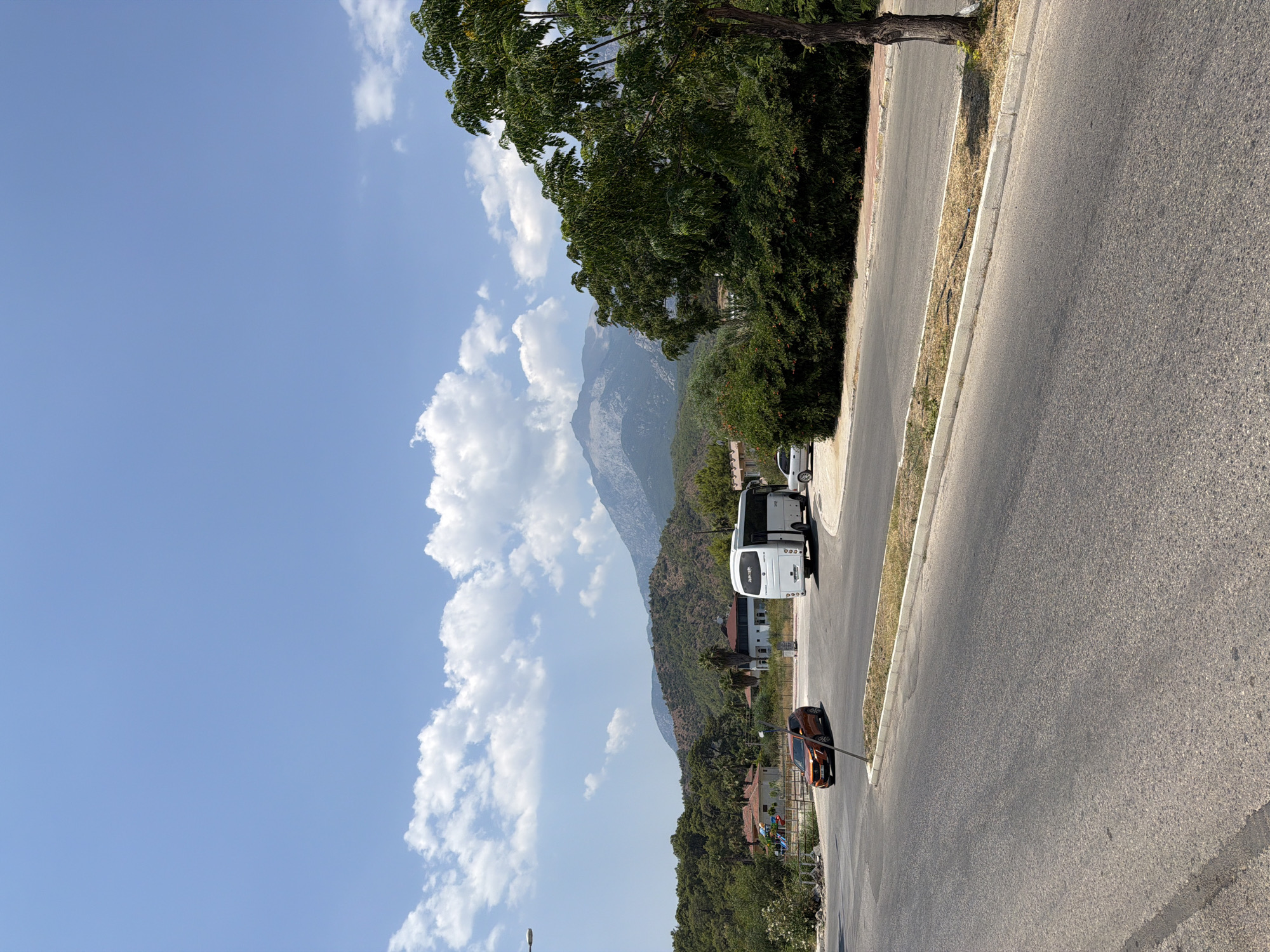 The image captures a scene in the city of Kemer, Turkey, on a clear, sunny day. In the foreground, a paved road with a white curb runs diagonally from the bottom left to the middle right. A light-colored car is parked on the side of the road. Further into the scene, a white bus is stopped, with its doors open. To the left of the bus, a small orange car is driving on the road. Behind the road and vehicles, there is a lush green area with trees and bushes. Beyond this, a series of hills covered in vegetation rise up, leading to a large, majestic mountain range in the background. The sky above is a bright blue, dotted with fluffy white clouds. The overall impression is a scenic and peaceful view of Kemer.