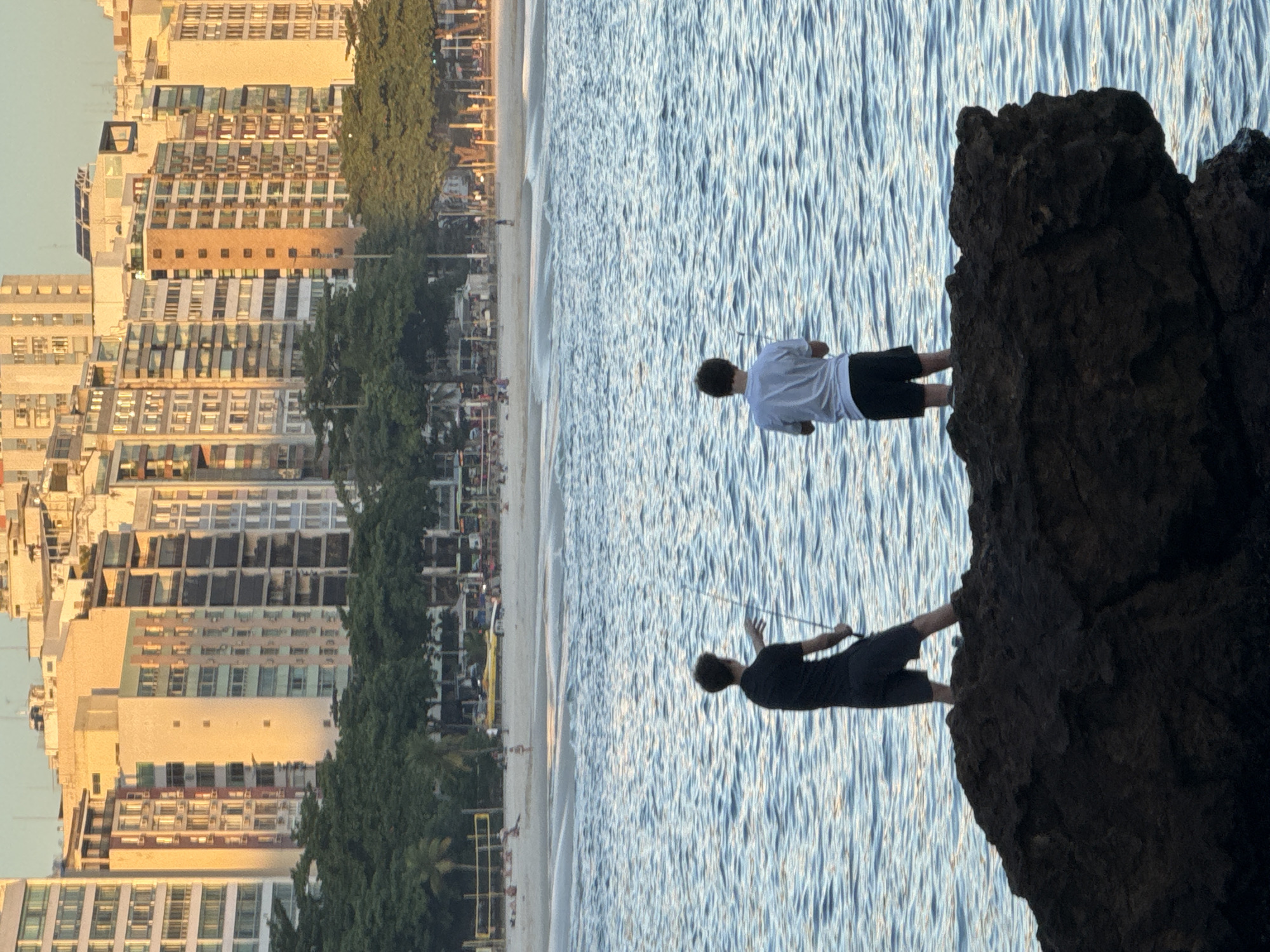 The image depicts a coastal scene in Niterói, Brazil, with a focus on two individuals fishing from a rocky outcrop. In the foreground, a dark, textured rock juts out into the calm blue water. Standing on the rock are two young males, viewed from behind. The individual on the left wears a black t-shirt and dark shorts, holding a fishing rod. To his right, the second individual is dressed in a white t-shirt and black shorts, also holding a fishing rod.
Beyond the water and the rock, a sandy beach is visible, with scattered people and beach umbrellas. Further inland, a line of lush green trees forms a boundary. Behind the trees, a dense collection of multi-story buildings stretches across the skyline, characteristic of a city. The architecture suggests an urban coastal environment. The lighting indicates it is either early morning or late afternoon, with warm sunlight casting a glow on the buildings. The ocean is relatively calm, with gentle ripples on the surface. The scene conveys a sense of leisure and outdoor activity within a city's waterfront.