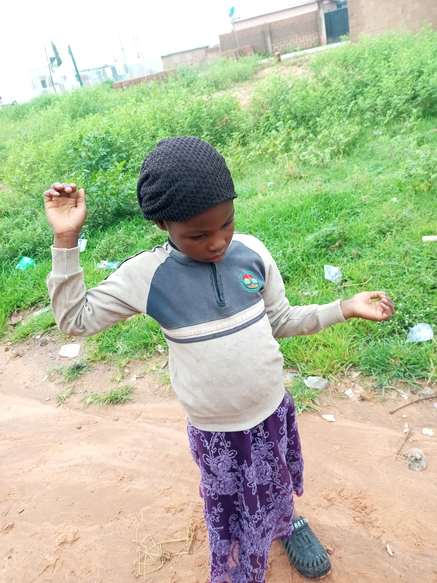 A young individual, likely a child, stands outdoors in a dusty, outdoor environment. They are wearing a dark, textured knitted cap, a light gray and dark blue long-sleeved shirt with a zipper at the collar and a circular emblem on the left chest, and a long, patterned purple skirt. Their right arm is extended outwards, and their left arm is bent upwards, with their left hand slightly open. The ground is a mix of reddish-brown dirt and sparse green grass. In the background, there is more overgrown green vegetation, and beyond that, some brick buildings and a pole with what appears to be a dish antenna. The overall atmosphere suggests a somewhat unkempt, possibly semi-rural or undeveloped area within Tempure, Nigeria.