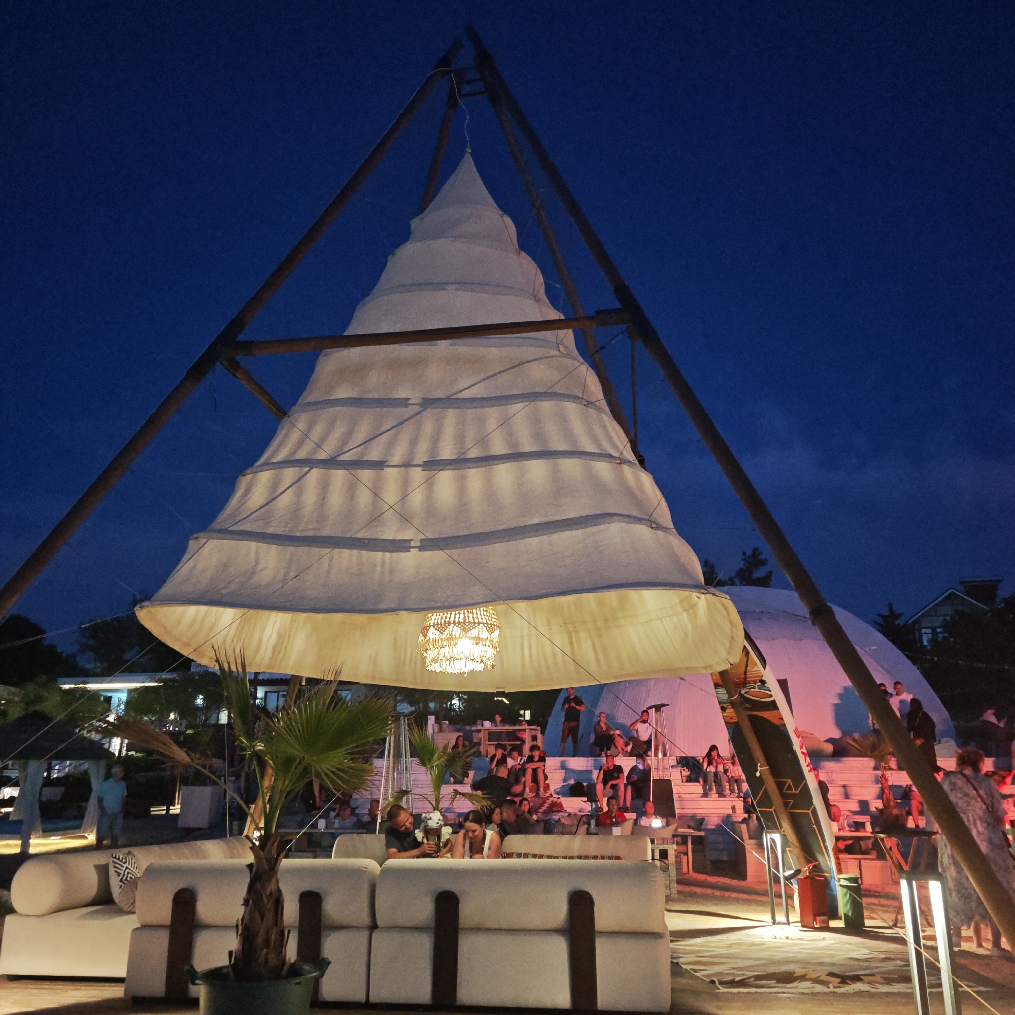 This is a medium shot of an outdoor evening event at a venue that appears to be on a beach or near water. In the foreground, there are several large, white modular couches. A small palm tree in a pot is situated to the left of the couches. A prominent feature of the scene is a large, conical-shaped structure made of white fabric, suspended from a wooden A-frame. A decorative chandelier hangs from the bottom of the fabric structure, illuminated by internal lights.
In the midground, a raised, tiered seating area is visible, with many people seated and standing. Some individuals are interacting with each other, while others appear to be watching an event or performance. There are also a few small tables and lights scattered throughout this area. In the background, there are buildings and trees, suggesting a developed area beyond the immediate venue. The sky is dark blue, indicating it is night or dusk. The overall atmosphere suggests a social gathering or entertainment event.