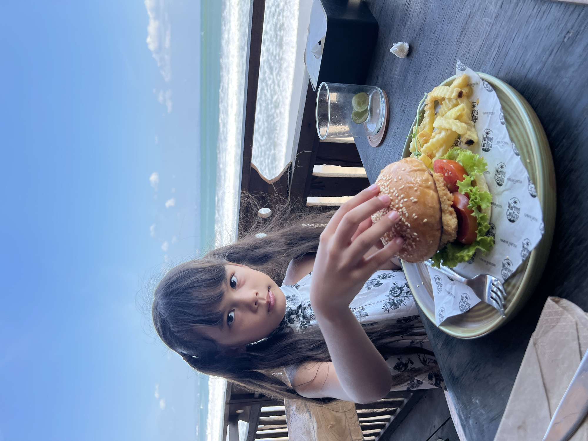 A young girl with dark hair in pigtails and a floral halter top is seated at a wooden table at a beachside restaurant in North Kuta, Indonesia. She is looking towards the camera with a neutral expression and reaching out to touch a hamburger on a plate. The hamburger is served with french fries, and there is a glass with lime slices on the table, along with a box of tissues. The background shows a wooden railing, a sandy beach, and the turquoise ocean under a clear blue sky with some scattered clouds. The restaurant appears to be open-air, with wooden structures and outdoor seating.