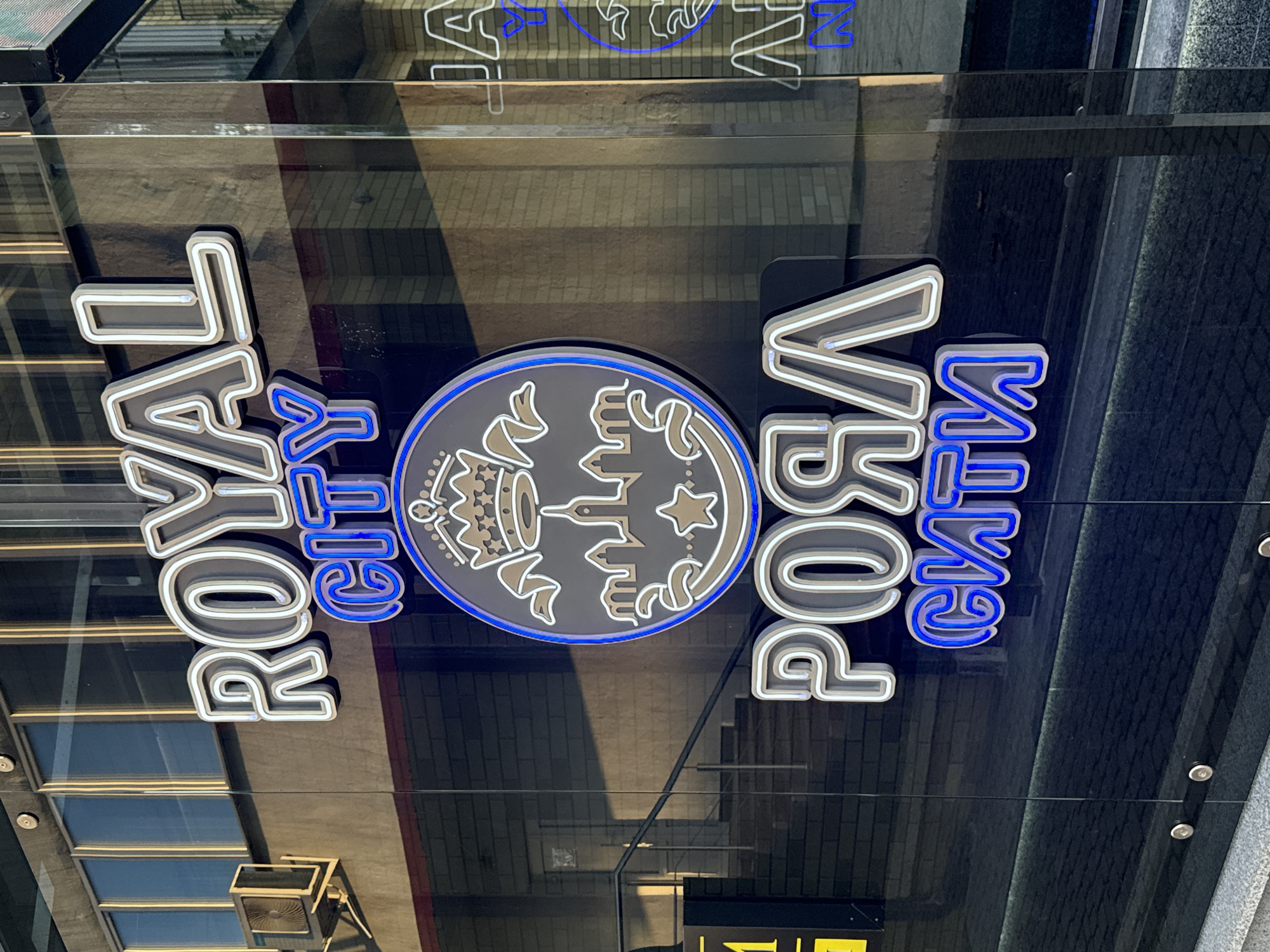 The image shows a storefront with neon signs. The main sign reads "ROYAL CITY" in large, block letters. Below this, a circular logo features a crown, a cityscape, and stars. Further down, the words "РОЯЛ СИТИ" are displayed in large, stylized letters, with "СИТИ" illuminated in bright blue neon. The storefront appears to be made of dark glass, reflecting the surrounding environment, including a brick building and a railing. An air conditioning unit is mounted on the exterior wall to the left of the sign. To the right, another neon sign is partially visible, with the letters "JA" and "N" illuminated in blue. The overall impression is of a business establishment, likely a club or entertainment venue, located in Mahilyow, Belarus.