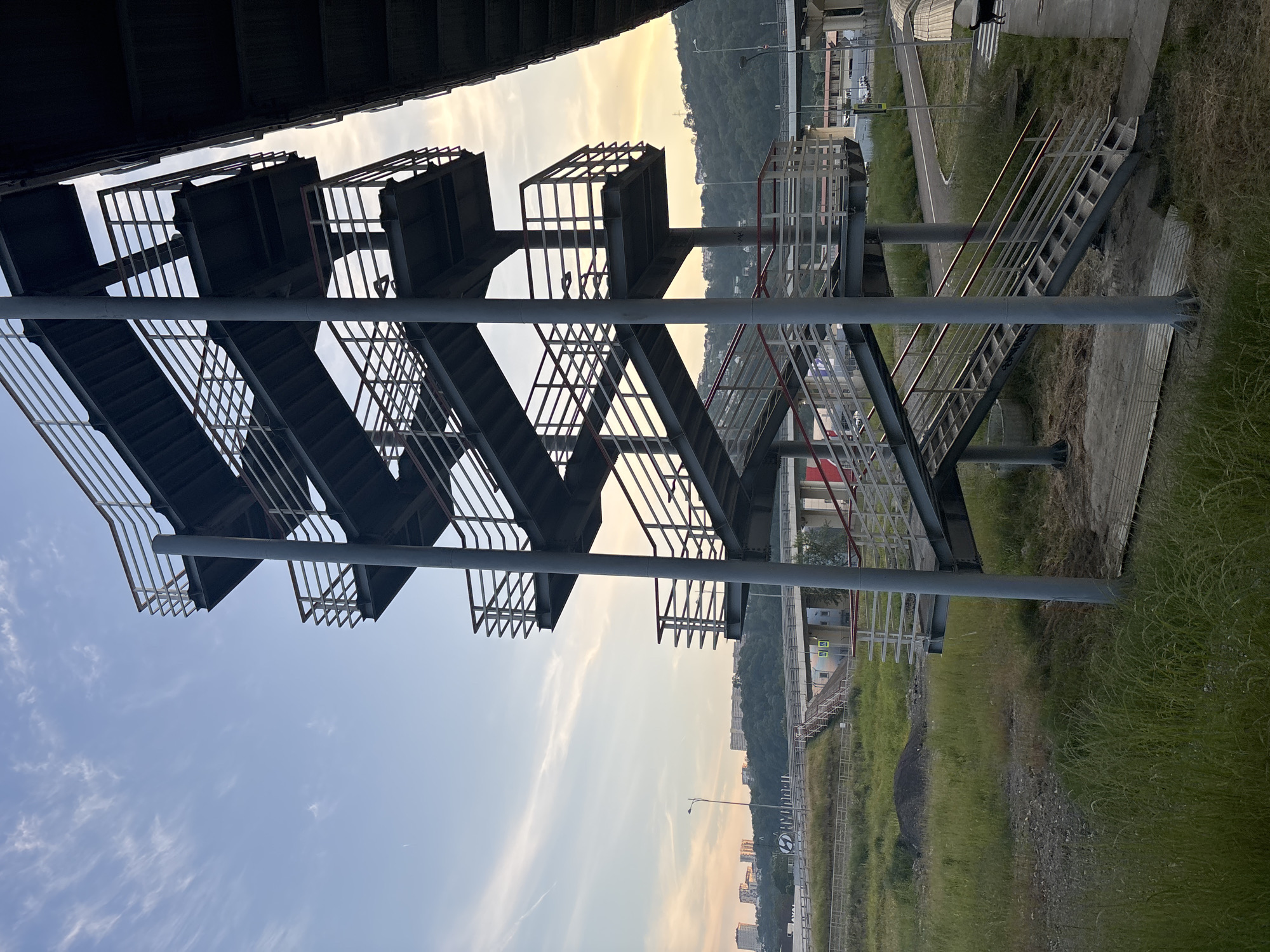 This image depicts a multi-tiered metal pedestrian overpass structure located in the Adler district of Sochi, Russia, specifically near Ulitsa Aviatsionnaya 34, crossing the A147 highway. The overpass is constructed from dark grey/silver metal, featuring multiple flights of stairs that ascend in a zig-zag pattern, connecting at least five visible square landings. The railings are a lighter grey with some red accents on the handrails, particularly in the lower sections. Tall, circular steel columns support the structure.
In the foreground, the ground is covered in unkempt green grass and weeds, interspersed with patches of bare earth and gravel. A small mound of dark material is visible to the middle-left. A concrete path or sidewalk with a partial crosswalk marking is present in the bottom right corner, where a small, dark-furred dog stands.
The midground features a complex multi-level highway interchange with several elevated roads, bridges, and underpasses. A prominent white sign bearing a blue "H" logo (Hyundai) is affixed to a bridge railing. A street light pole stands near the overpass. Beyond the road infrastructure, hills covered in dense green trees rise in the background, with several modern multi-story apartment buildings visible on their distant crests.
The sky transitions from a clear light blue at the top to a warm orange and pink hue near the horizon, indicative of late afternoon or early evening. Scattered wispy white cirrus clouds and a faint airplane contrail are visible in the upper-left sky. No people are present in the scene.