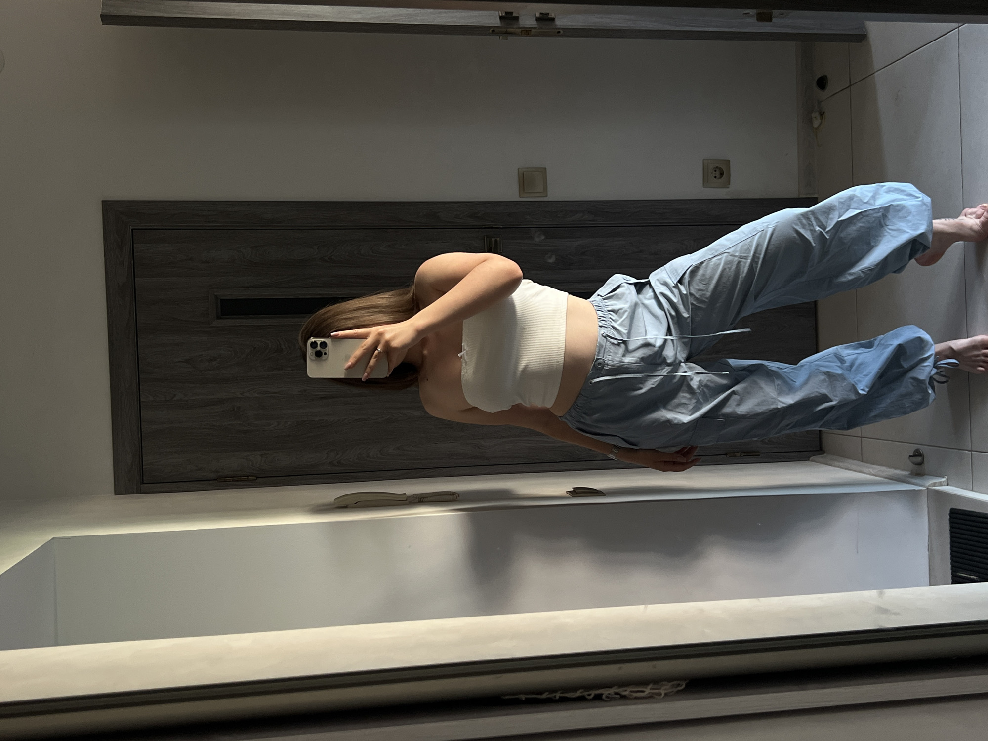 A young woman is posing for a mirror selfie in what appears to be an indoor setting, likely a bathroom or dressing area, in Ternopil, Ukraine. She is wearing a white ribbed tube top and light blue baggy cargo pants. Her right hand is holding a smartphone, angled to capture her reflection. Her left arm is bent and raised, with her hand resting on her head, obscuring part of her face. She has long, light brown hair. The background shows a modern interior with a dark wooden door, a wall with a light switch and an electrical outlet, and a large mirror reflecting the scene. The overall impression is casual and everyday. There is no visible text in the image.