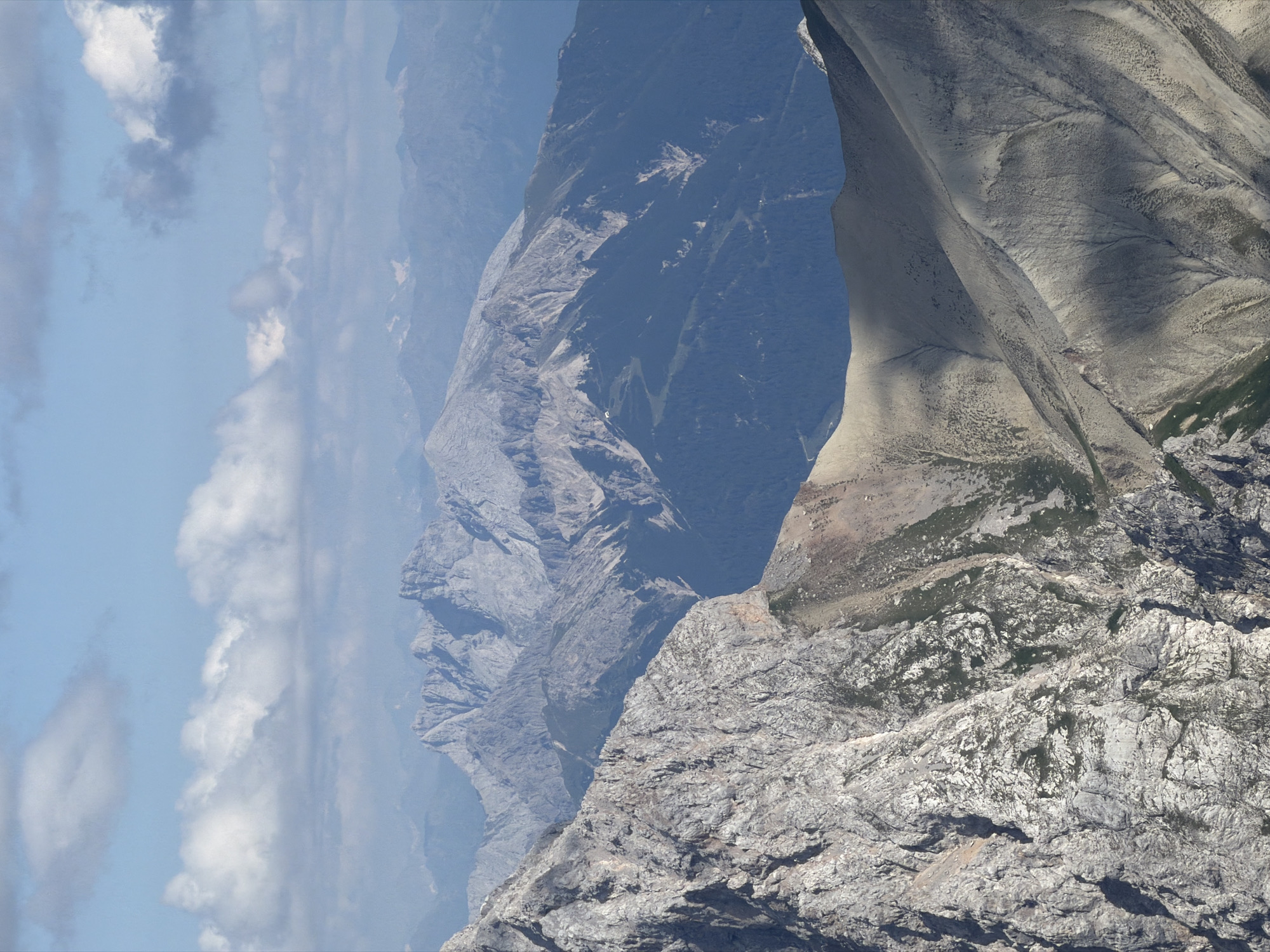 This is a photograph taken from a high vantage point, looking out over a vast mountain range. The foreground features a rugged, rocky mountain slope, with sharp, jagged peaks and crevices. The texture of the rock is detailed, showing variations in color from light gray to darker shades, with patches of green vegetation clinging to some surfaces.
In the middle ground, a series of progressively distant mountain ranges are visible. The closer ranges are covered in dark green forests, while the further ranges are lighter shades of gray and blue, fading into the distance. The terrain is characterized by steep slopes, deep valleys, and prominent peaks.
The background consists of a hazy, blue sky with scattered white clouds. The overall impression is one of immense scale and natural beauty, showcasing the grandeur of the Alps. The scene is likely captured during daylight, with the sun casting shadows across the mountains, highlighting their contours.
The photograph is taken from a high altitude, suggesting the photographer is on another mountain or a very high point. The specific location in Garmisch-Partenkirchen, Germany, is not explicitly evident from the image itself, as it depicts a natural, mountainous landscape. However, such a vista is consistent with the scenery found in the Bavarian Alps surrounding Garmisch-Partenkirchen. There are no people, animals, or man-made structures directly visible within the frame, focusing solely on the natural environment.