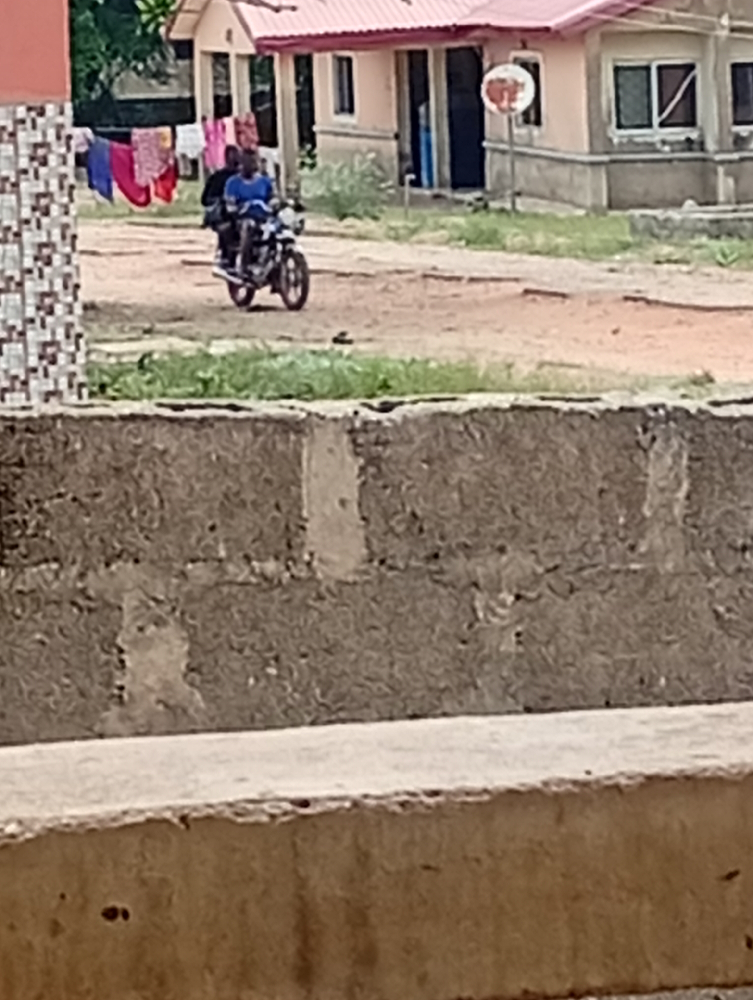 The image captures an outdoor scene, likely a residential area in Adankolo, Nigeria. In the midground, two individuals are seen riding a dark-colored motorcycle away from the viewer. The person driving is wearing a blue shirt.
Behind the motorcycle, several simple, single-story or low-rise buildings are visible under what appears to be clear daylight. A prominent building features a distinctive red-pink roof and light-colored walls, possibly painted concrete. A small, round, discolored satellite dish is mounted on a pole near this building. Between buildings on the left, various pieces of laundry, including blue, red, and pink garments, hang on a clothesline. The ground beneath the motorcycle and leading up to the buildings is an unpaved, brownish dirt road with sparse patches of green vegetation.
The foreground is dominated by a rough-textured, dark brown concrete wall or foundation, which spans the width of the frame. In front of this, a lighter-colored, smoother concrete slab is visible. No discernible text is present in the image. The overall impression is that of a typical street or pathway in a developing community.