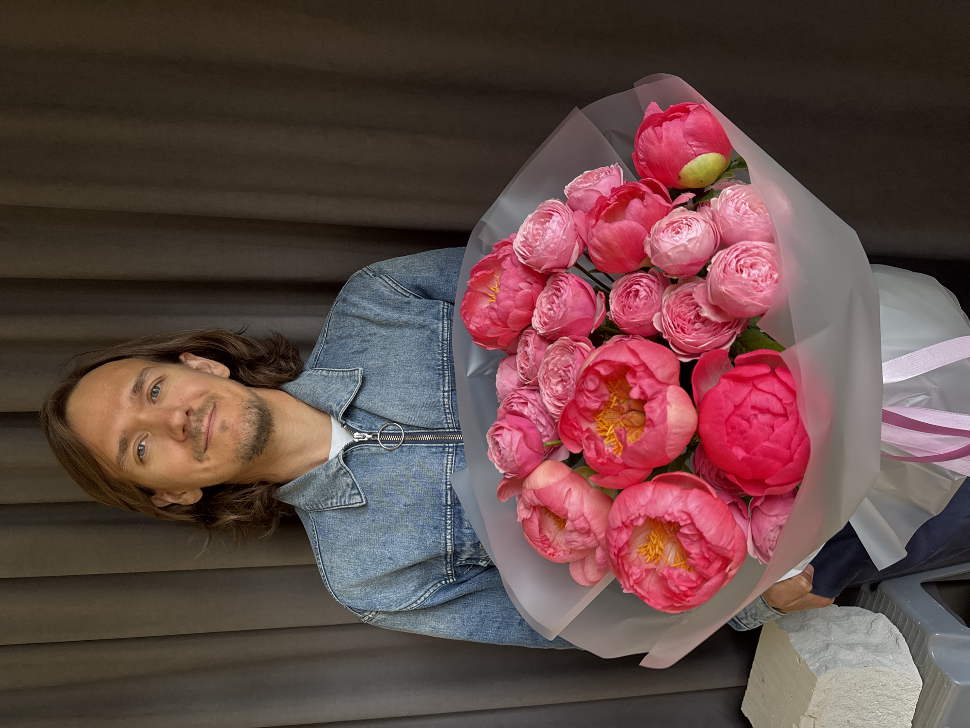 A man is holding a bouquet of pink peonies wrapped in translucent paper with a pink ribbon. He is wearing a denim jacket over a white shirt. The background is a brown curtain. The scene appears to be outdoors, with a piece of gray stone visible at the bottom right. The photo was taken in the city of Izhevsk, Russia.