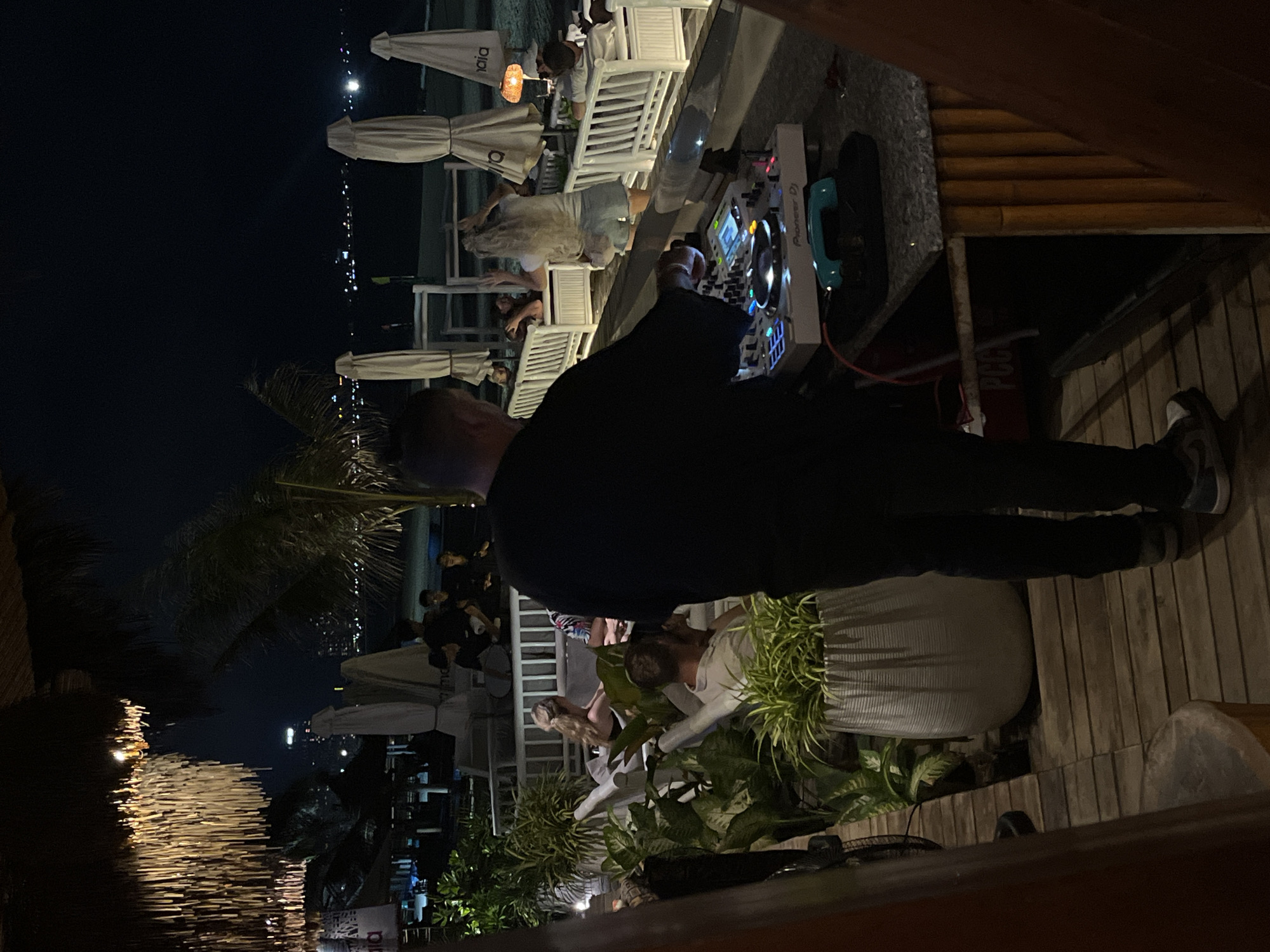 The photograph captures a nighttime scene in Đà Nẵng, Vietnam, likely at a beachside establishment. A DJ, dressed in dark clothing, is actively using a Pioneer DJ set, indicating a live performance. Several patrons are seated on white patio furniture under parasols. The outdoor venue is decorated with plants, including large potted shrubs near the wooden deck where the DJ is positioned. The city lights are visible in the distance, suggesting a coastal location. A partially visible logo for "PCC" can be observed on a red object under the DJ's setup.