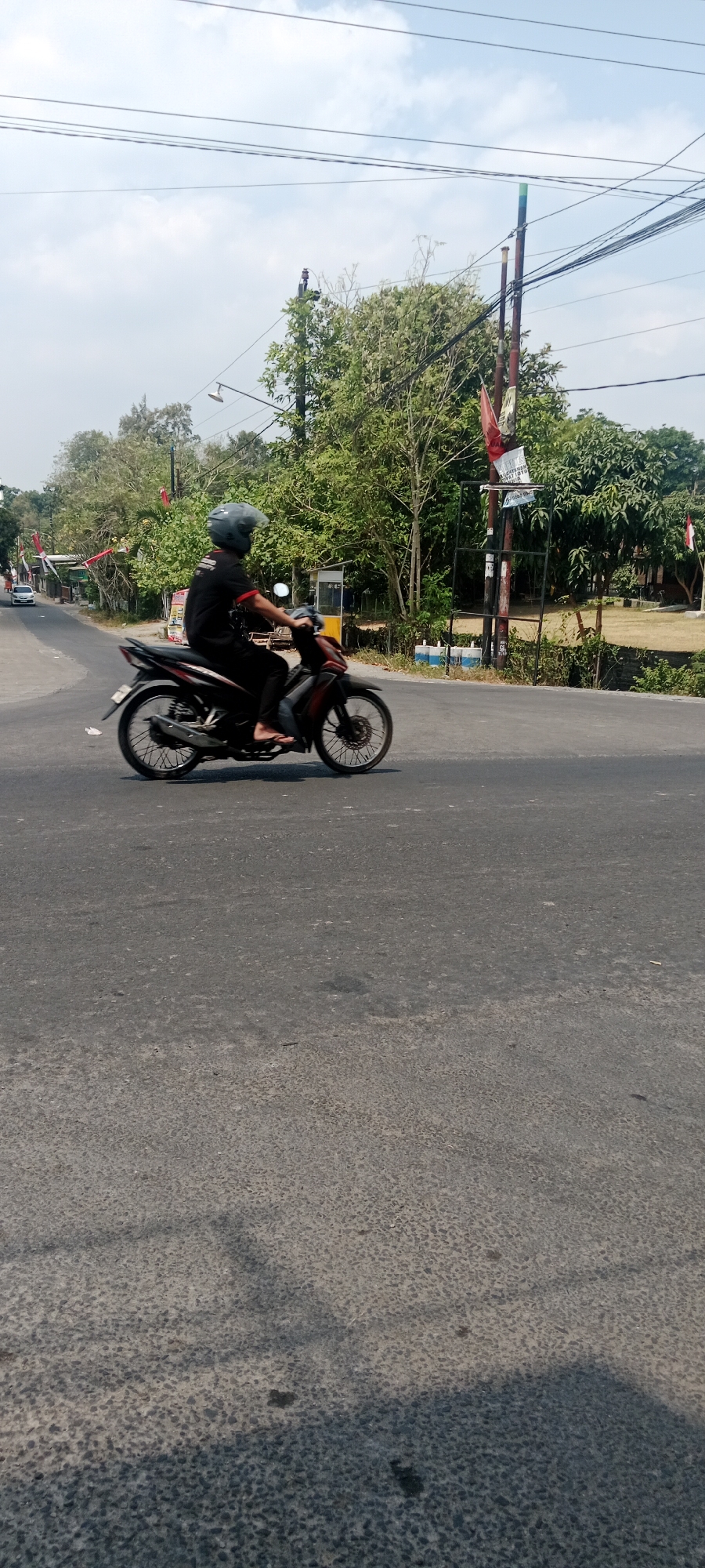 A person wearing a grey helmet and a black t-shirt is riding a dark-colored motorcycle on an asphalt road in Maguwoharjo, Indonesia. The motorcycle has red and black accents and spoked wheels. The rider is wearing flip-flops. The road is a two-lane street with a slight curve. Trees and some buildings are visible on the left side of the road, with a white car in the distance. On the right side of the road, there are utility poles with wires, some flags, and more trees. The sky is partly cloudy. The overall scene depicts a typical day on a road in a semi-urban or suburban area.