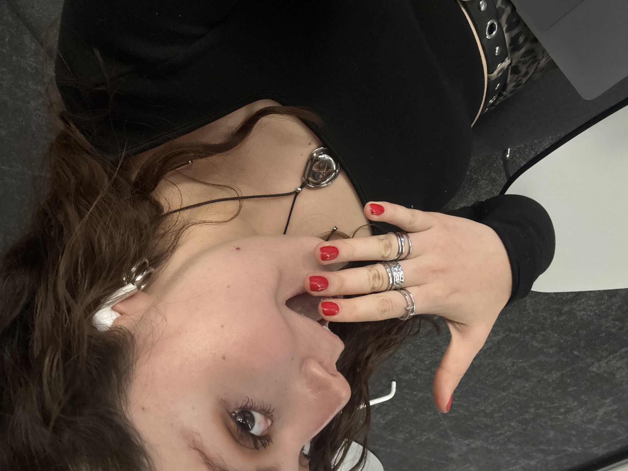 A woman lies on a gray carpeted floor. She is wearing a black long-sleeved top and a belt with silver grommets. She has long, curly brown hair and is wearing a silver necklace and earrings. She is also wearing several silver rings on her right hand, which she has raised to her mouth. Her fingernails are painted red. A white table and a laptop are visible in the upper right corner of the image. The location is unknown and there is no evidence to indicate that it is in Mexico City, Mexico.