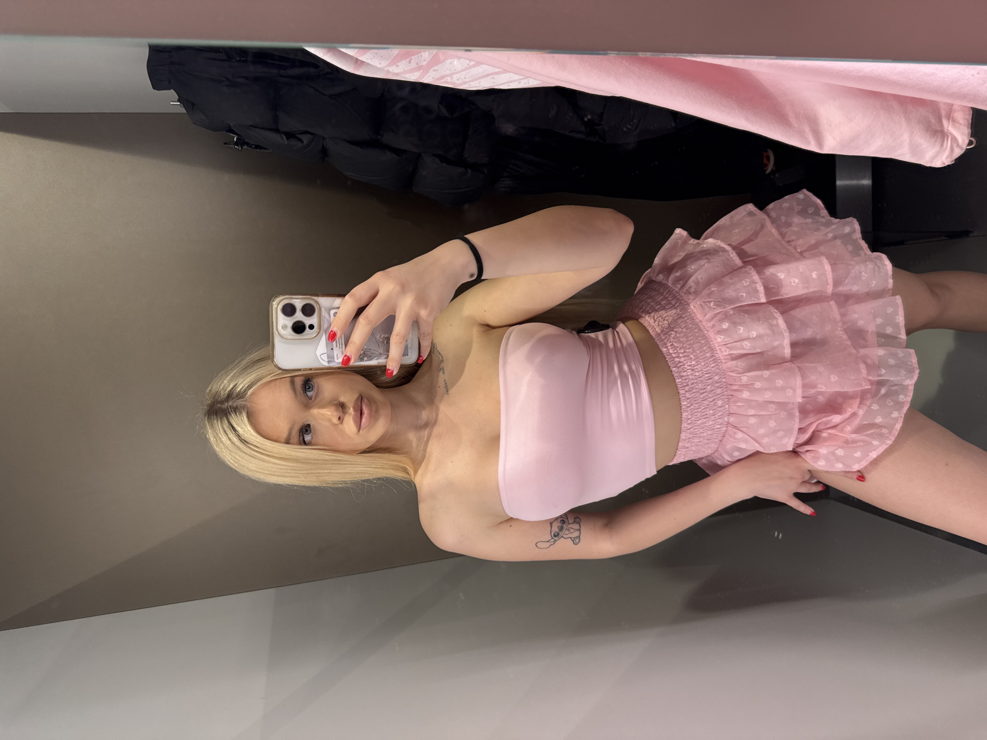 A young woman with blonde hair is taking a mirror selfie in what appears to be a fitting room or dressing area. She is wearing a pink tube top and a pink ruffled skirt with heart patterns. Her left arm has a small tattoo of the character Stitch. She is holding a smartphone in her right hand, which has red nail polish. In the background, there are garments hanging on hooks, including a black puffer jacket and a pink piece of clothing. The room has grey and brown walls and a large mirror. The location is specified as Kudrovo, Russia.