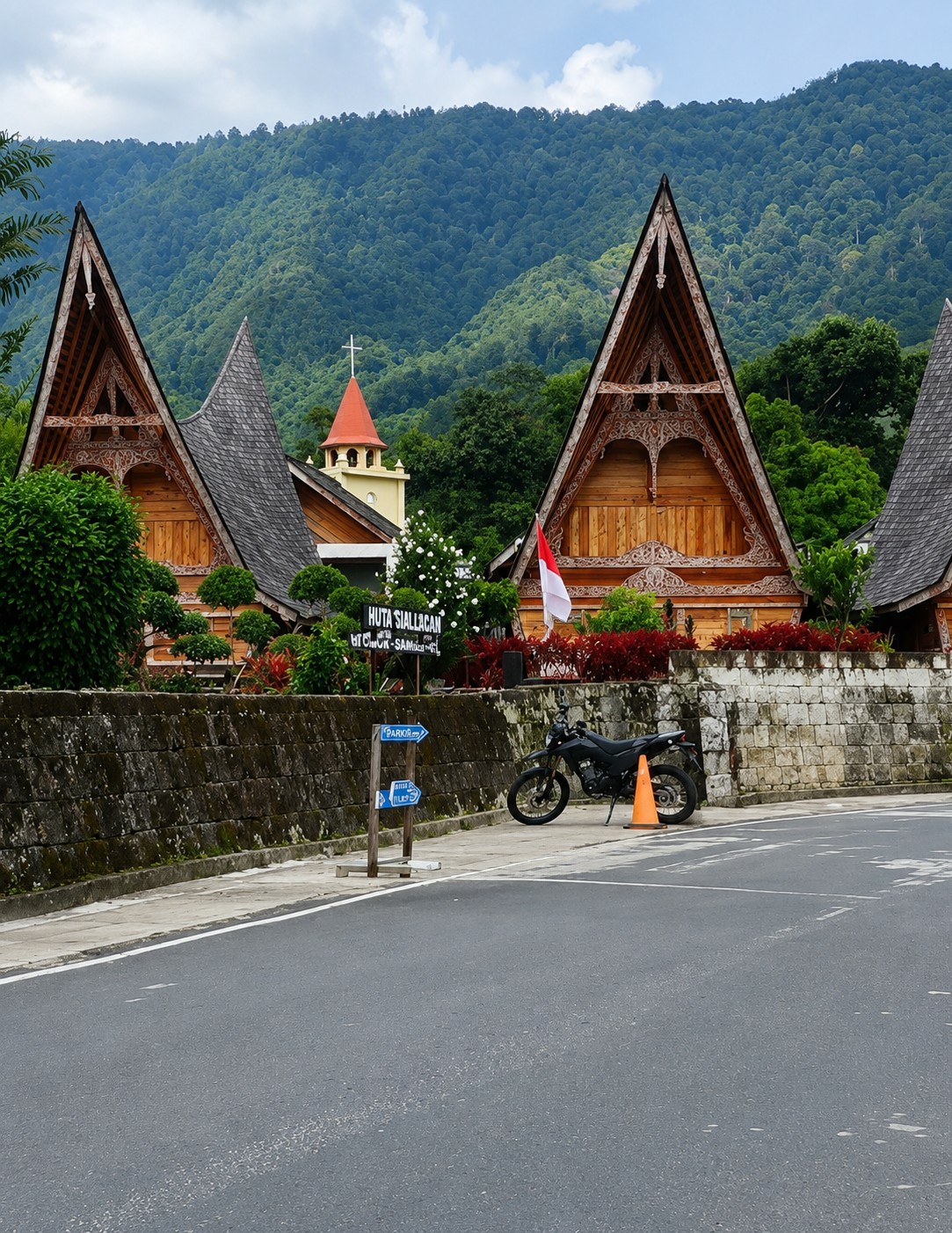 The image captures a scenic view in Ambarita, Indonesia, featuring traditional Batak architecture set against a lush, forested mountain backdrop. In the foreground, a paved road runs alongside a low stone wall. A black motorcycle is parked on the sidewalk next to the wall, with an orange traffic cone beside it. Two blue directional signs are mounted on wooden posts near the motorcycle, indicating "PARKING" to the left and an arrow pointing to the right. Behind the wall, several wooden houses with distinctive triangular roofs, adorned with intricate carvings, are visible. The Indonesian flag is displayed on one of the houses. A church steeple with a red roof and a white cross rises above the houses. The surrounding area is filled with greenery, including trees and red foliage. The sky is partly cloudy, and the mountains in the distance are covered in dense forest.