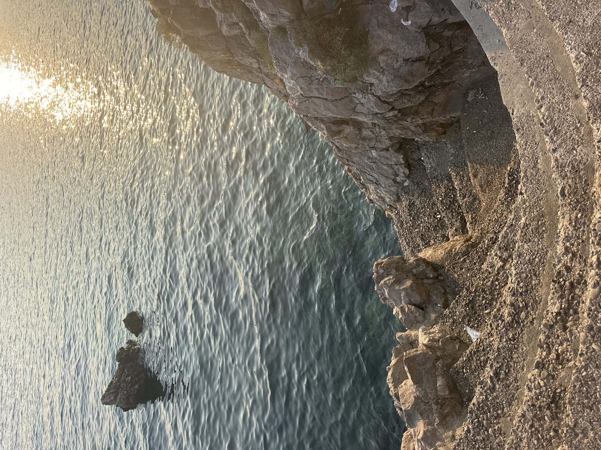 A coastal scene in Budva, Montenegro, features choppy, dark blue ocean water reflecting the golden sunlight on the right side of the frame. Two dark rocks jut out of the water in the upper left quadrant. The foreground is dominated by a rocky, uneven shoreline with some scattered pebbles and larger rocks. A narrow, worn concrete pathway or set of steps curves from the lower right corner upwards towards the rocky cliff face. This cliff is rugged and textured, with sparse green vegetation clinging to its surface. Scattered white litter, including what appears to be cigarette butts and a plastic bottle, is visible on the ground and near the base of the cliff. The overall impression is of a natural, somewhat unkempt seaside location.