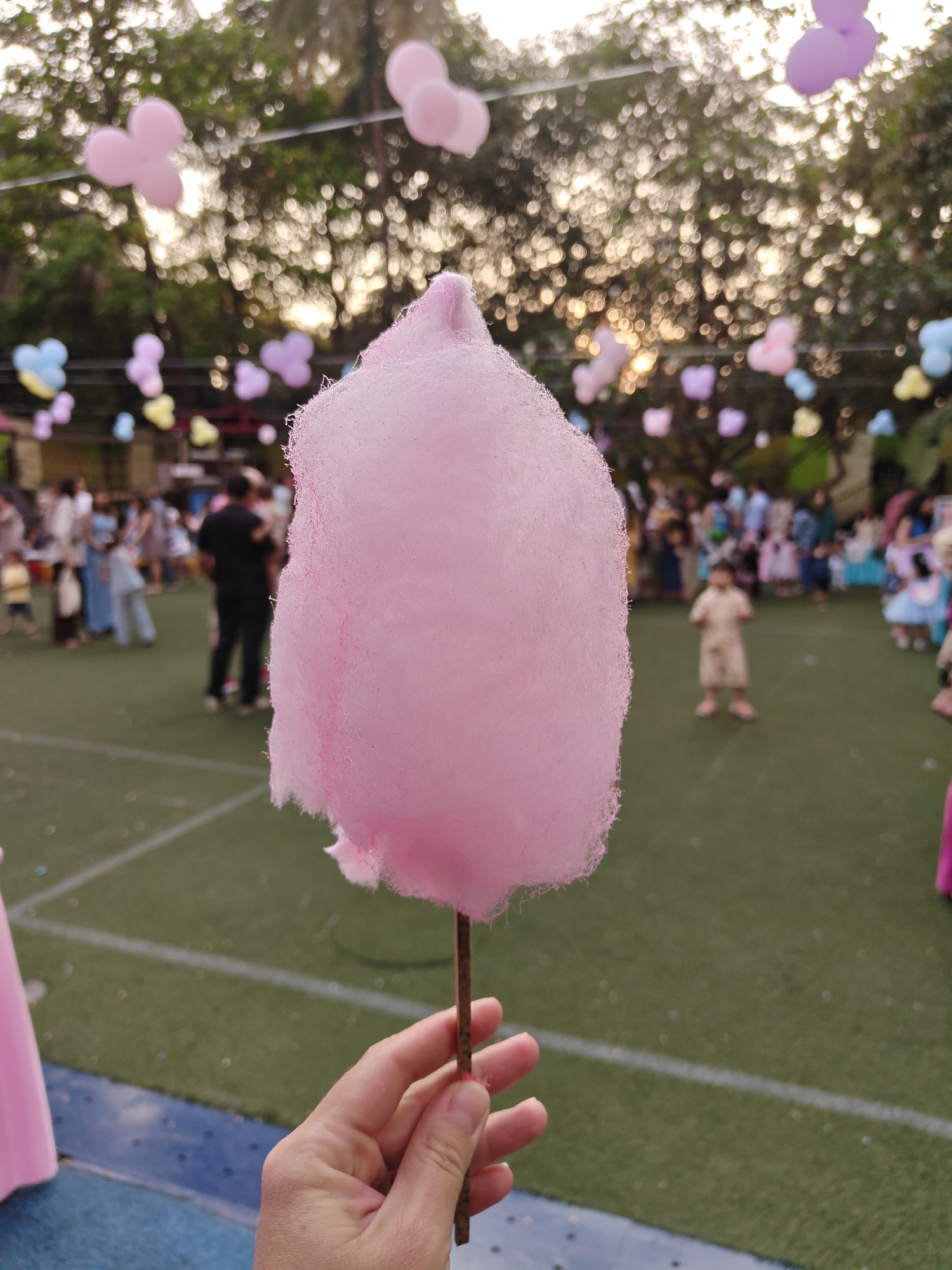 A close-up, first-person perspective shows a fair-skinned human hand holding a stick of pink cotton candy, centrally positioned and in sharp focus. The background, extensively blurred, reveals an outdoor gathering taking place on a green artificial turf surface, marked with white lines, likely in Mumbai, India. Numerous indistinct figures, comprising adults and children in varying attire, are present across the mid-ground and background. Overhead, multiple clusters of balloons or similar decorative items in pink, purple, yellow, and blue are suspended. Lush green trees are visible in the distant background, illuminated by a warm, diffused light suggesting late afternoon or early evening, creating prominent bokeh effects. A blue border is visible at the bottom left edge of the turf.