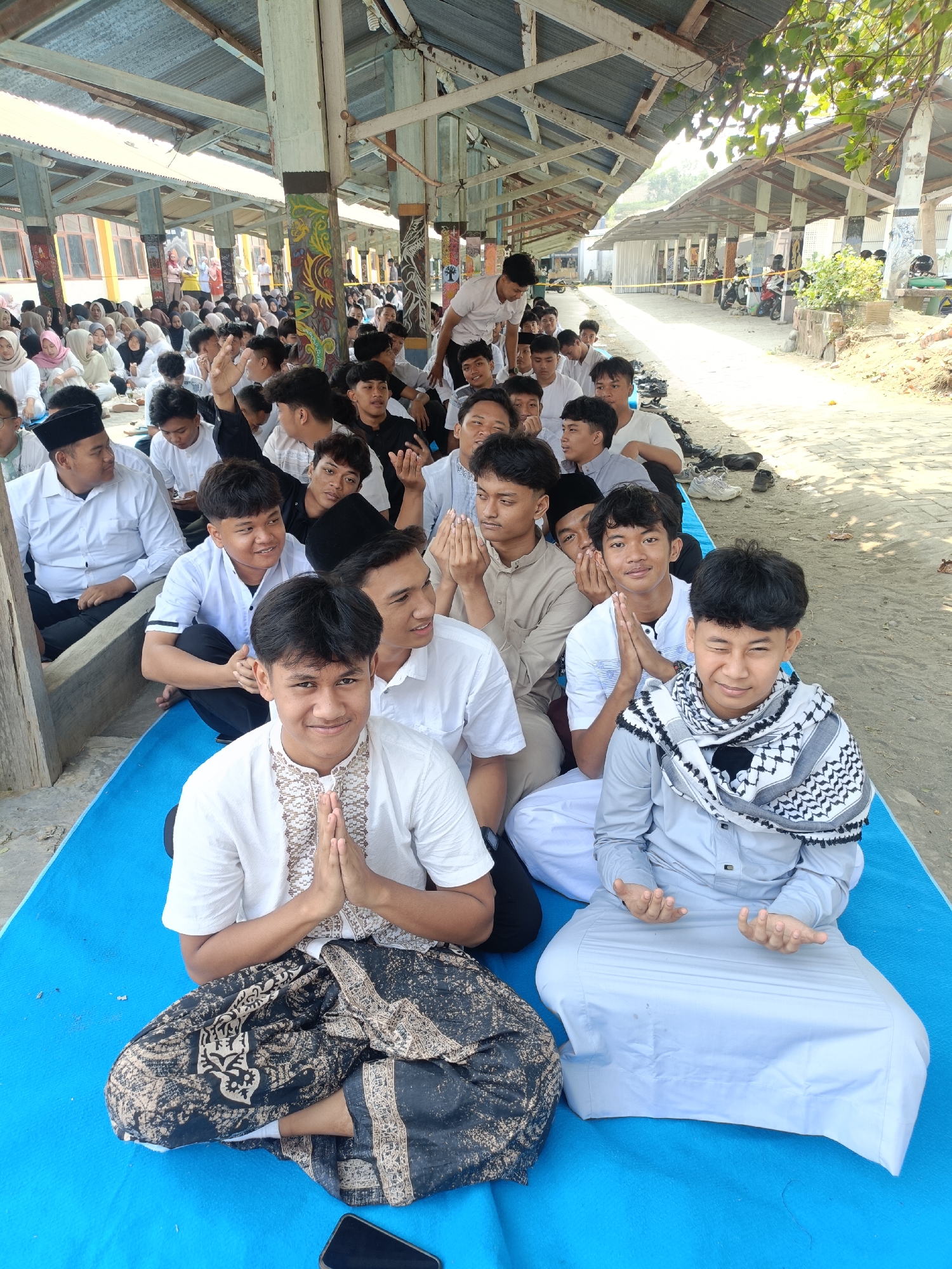 A group of young Indonesian men in Islamic attire are seated on a blue mat outdoors under a tin roof structure. Some are looking at the camera, and one is smiling and winking. Others have their hands pressed together in prayer. In the background, more people are seated and standing, with a building visible. The scene appears to be in Kediri City, Indonesia, likely a religious gathering.