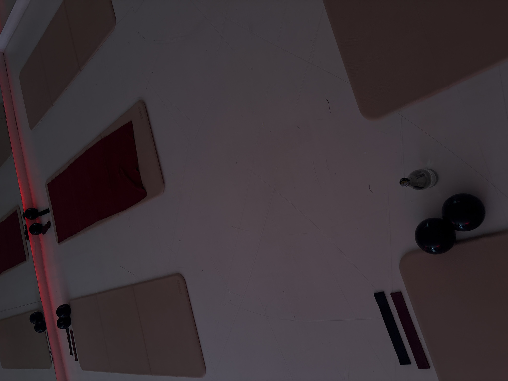 The image depicts an interior scene of a fitness or wellness studio located in Mexico City, Mexico. The environment is a dimly lit room with a light-colored floor, possibly white or light grey, exhibiting numerous fine scratches or scuff marks. A mirrored wall spans the upper portion of the frame, with a horizontal red LED light strip positioned along its base, casting a red glow into the space.
Multiple pieces of exercise equipment are arranged on the floor. Four rectangular mats are visible: one partially cropped in the lower right foreground, one centrally positioned and covered with a dark red fabric, and two light beige mats positioned to the left and far right. On the light beige mat in the mid-ground left, two black spherical objects, likely small weights or massage balls, are present alongside two dark, flat, rectangular items, possibly resistance bands. Similar items are located on the floor in the lower-left foreground next to a partially visible light beige mat: two black spherical objects and two dark rectangular bands. A clear glass bottle with a silver cap is situated on the floor near the bottom-center, to the right of the spherical objects. Reflections of equipment, including black spherical and handle-shaped objects, are visible in the mirror above the dark red mat. No individuals are present in the scene.