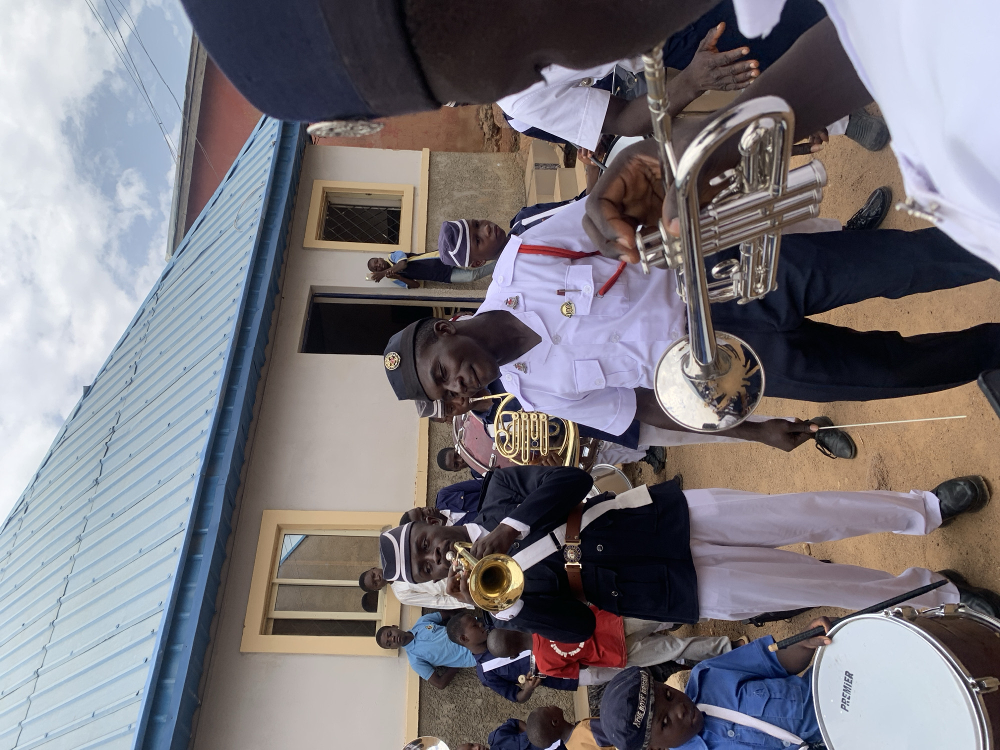 A ground-level, slightly canted, medium shot depicts a marching band on dirt near a building with a corrugated blue roof, likely in Jos, Nigeria. The foreground features several people in marching band uniforms, including navy blazers and white pants with dark shoes. A person in a white shirt with a red strap is holding a silver trumpet towards the camera's left. Another band member in the lower left is holding a brass trumpet and wearing a navy blazer and white pants. At the bottom-right, a drummer with a blue uniform and cap is holding a drumstick near a "Premier" drum. In the background, other band members are visible, including one with a tuba and others wearing similar uniforms. The building with the blue roof has light beige walls and a few visible windows. People are looking out of the window. The overall atmosphere appears to be that of a band performance or practice.
