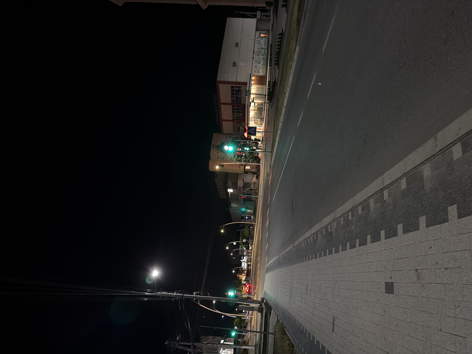 The image depicts a night-time street scene in Strășeni, Moldova. A paved road with white lane markings is visible, with a sidewalk made of light and dark patterned tiles running along the left side. Streetlights illuminate the area, casting a glow on the road and surrounding buildings. Traffic lights are visible, with some showing green. A red car is crossing the intersection in the distance. Buildings line the street, including what appears to be a supermarket with a sign that reads "SUPERMARKET". There are no people clearly visible in the image, and no discernible actions or interactions are taking place. The overall environment suggests a typical urban street at night.