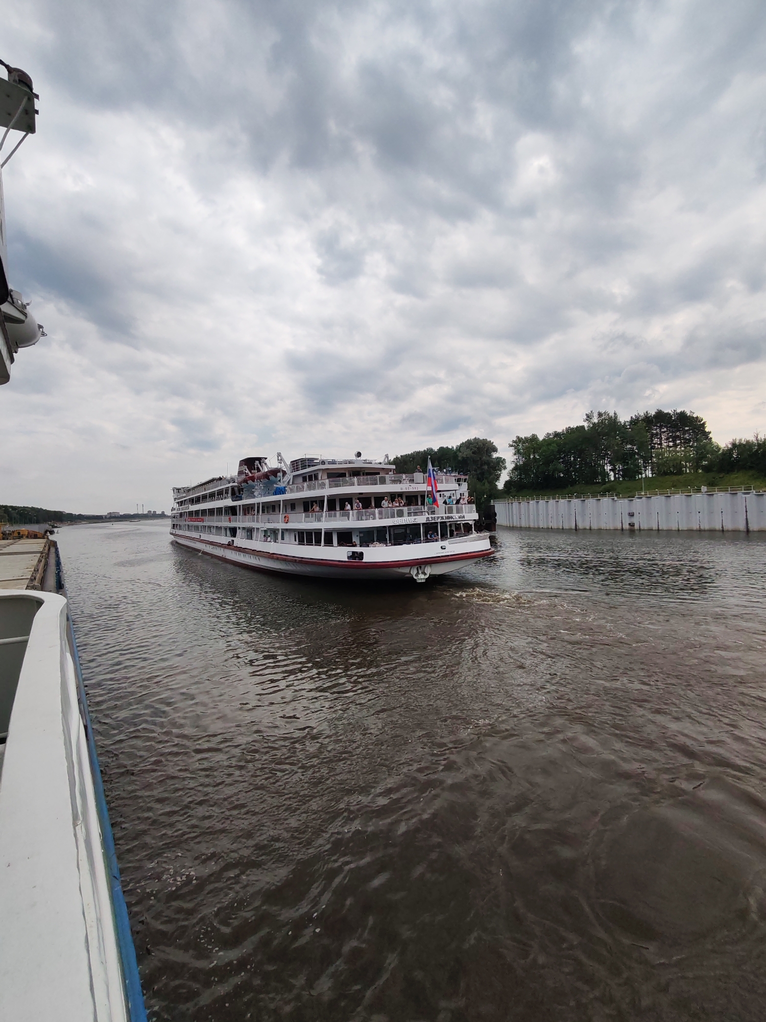 This is a full-frame, eye-level, wide-angle, outdoor, natural-light photograph taken from a boat. The scene depicts a large, white passenger ship with multiple decks, adorned with red trim and several flags, moving through a river. The ship is in motion, creating a wake in the dark, rippled water. A portion of another boat, also white with blue trim, is visible in the foreground on the left side of the frame.
In the background, on the right side of the river, a concrete wall or embankment runs along the bank, topped by a line of dense green trees. The sky above is overcast with dramatic, gray clouds. The overall environment suggests a waterway within or near a city, possibly during daytime with an approaching storm or a generally cloudy day.
The ship appears to be a passenger vessel, likely a river cruise ship, with people visible on its decks, observing the surroundings. The name "ДЗЕРЖИНСКИЙ" is visible on the side of the ship, which is a proper noun, possibly the ship's name. The image is taken on the river in the settlement of Bol'shie Mkhi, Russia.
