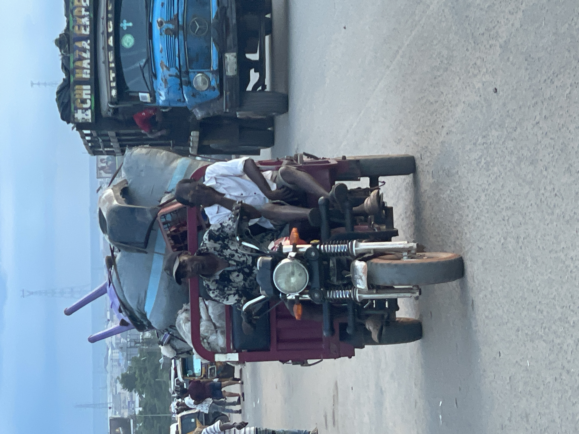 In Onitsha, Nigeria, on a paved road under a cloudy sky, two men are riding in a maroon-colored three-wheeled vehicle laden with bulky cargo, including what appears to be large purple-tipped plastic objects and gray tarps. The driver, wearing a patterned shirt and a black cap, is operating the vehicle. Beside him, a passenger in a white shirt is holding his hand to his face. Behind them, a blue truck with "CHI HAZA EXPRES" written on its side is visible. The scene depicts a busy urban environment with other vehicles and pedestrians in the background, suggesting a bustling marketplace or transportation hub.