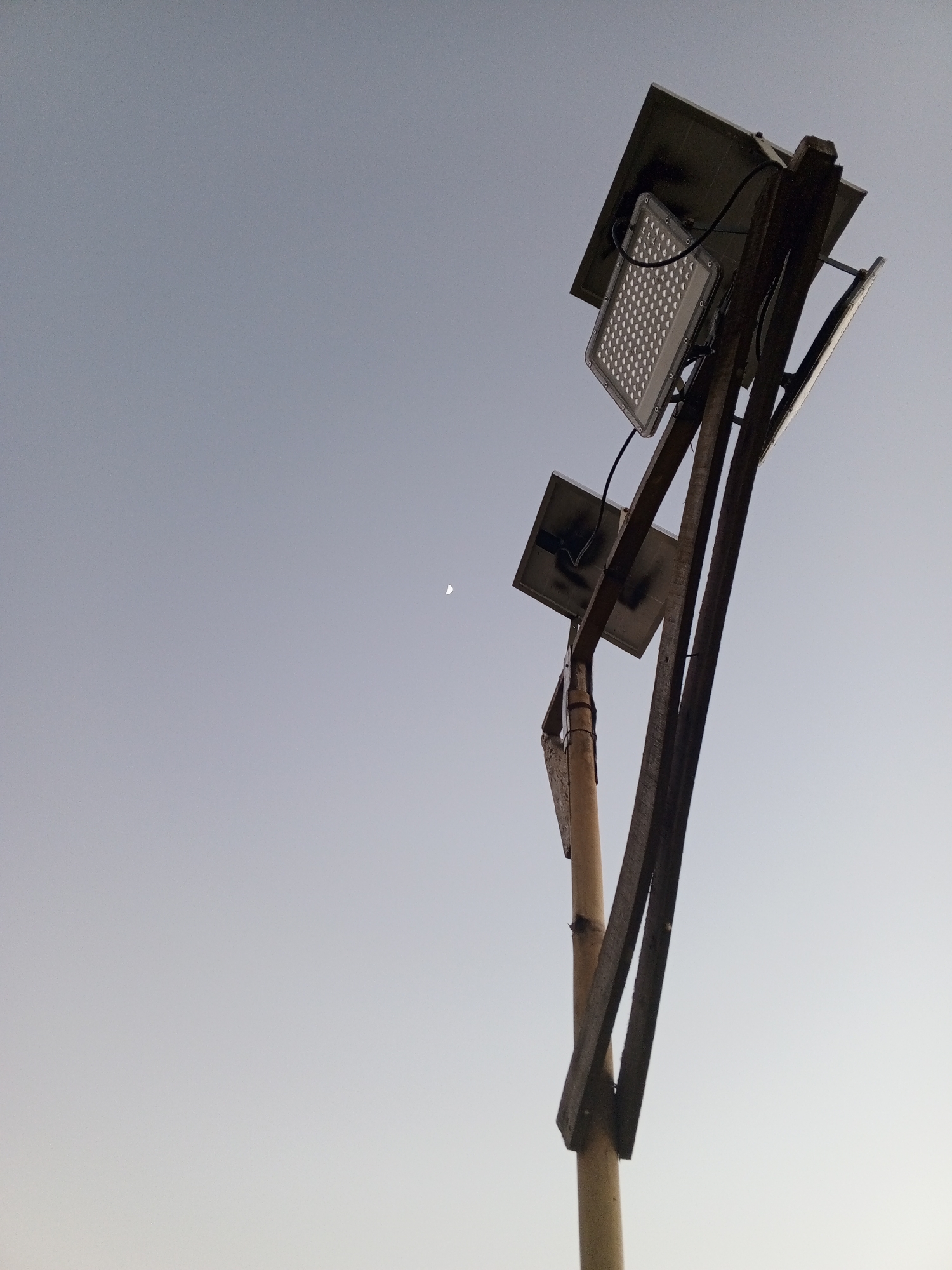A low-angle shot captures a solar-powered street lamp against a pale sky, with the crescent moon faintly visible in the distance. The lamp fixture, which appears to be an LED floodlight, is mounted on a wooden pole with additional solar panels. The structure is rustic, with visible wires connecting the components. The scene is devoid of any people or animals, creating a serene yet functional aesthetic. The location is Watagoda, Sri Lanka.