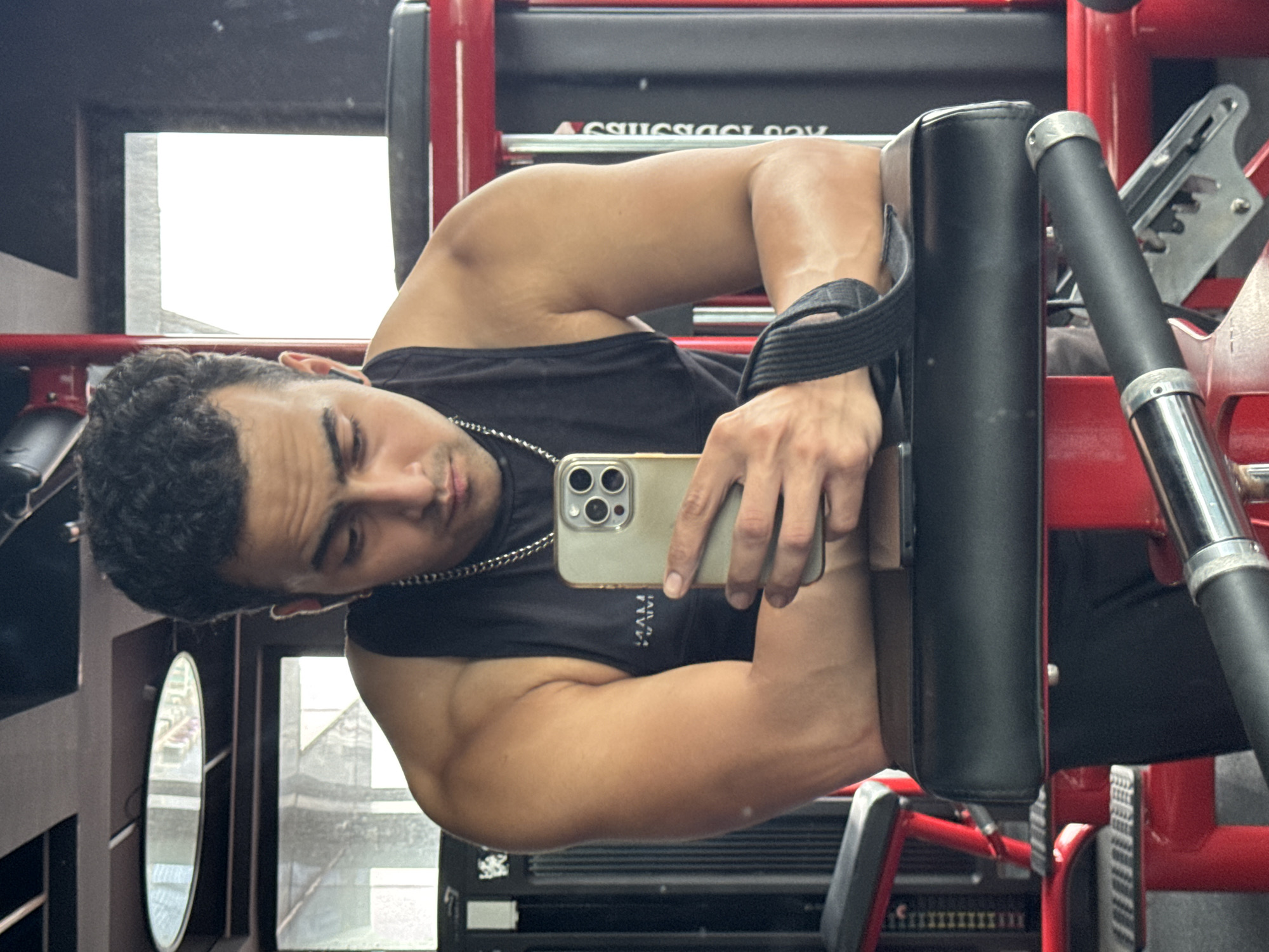 A South Asian man with dark, curly hair and a light brown complexion is taking a selfie in a gym. He is wearing a black sleeveless t-shirt, a silver chain necklace, and wireless earbuds. His right arm rests on a black padded equipment seat, and his left hand holds a light gold smartphone. A black strap is wrapped around his left wrist, attached to the equipment. The gym equipment is red and black, and the background shows mirrors and windows. The image does not provide any information about the specific location in Kolkata, India.