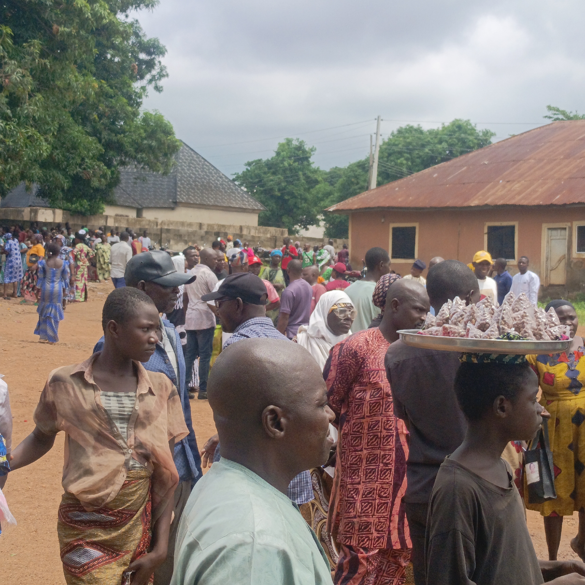 The image depicts a crowded outdoor scene in Akwanga, Nigeria, likely at a social gathering or market. The ground is dirt. There are a few buildings in the background, with a red metal roof and a grey tiled roof. Many people are dressed in patterned clothing. A vendor is carrying goods on their head. There is a woman wearing a headscarf and eyeglasses. A boy is carrying a tray with many small plastic bags containing something.