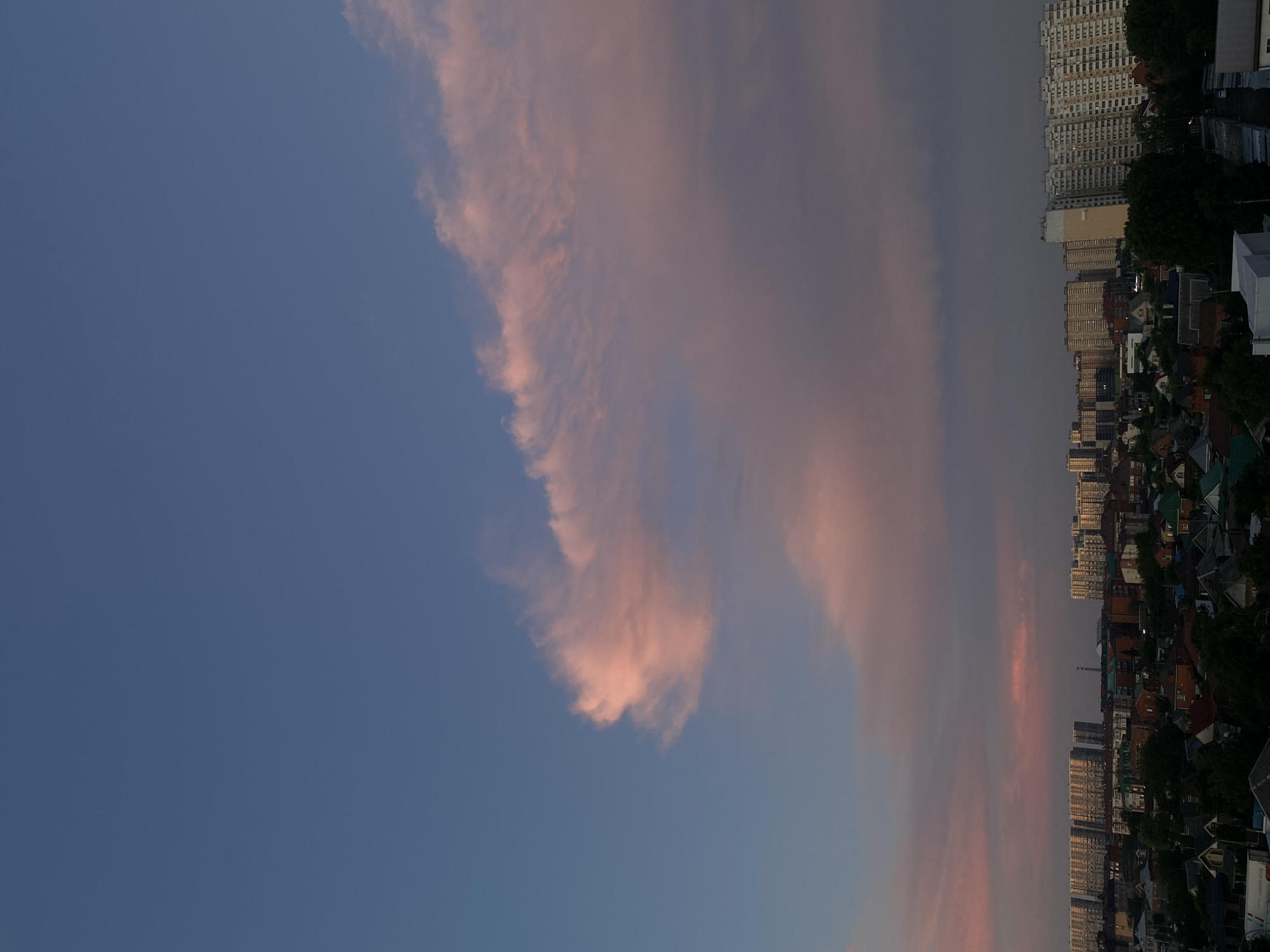 The image shows a cityscape with a dusky sky. In the foreground, there is a dense area of houses with pitched roofs, some of which are red or green. Beyond the houses, there are several multi-story apartment buildings, some of which are tall and rectangular, while others are shorter and more varied in design. The sky is a deep blue at the top, transitioning to shades of pink and purple lower down, with large, wispy clouds. The lighting suggests it is either sunset or sunrise. There are no people visible in the image.