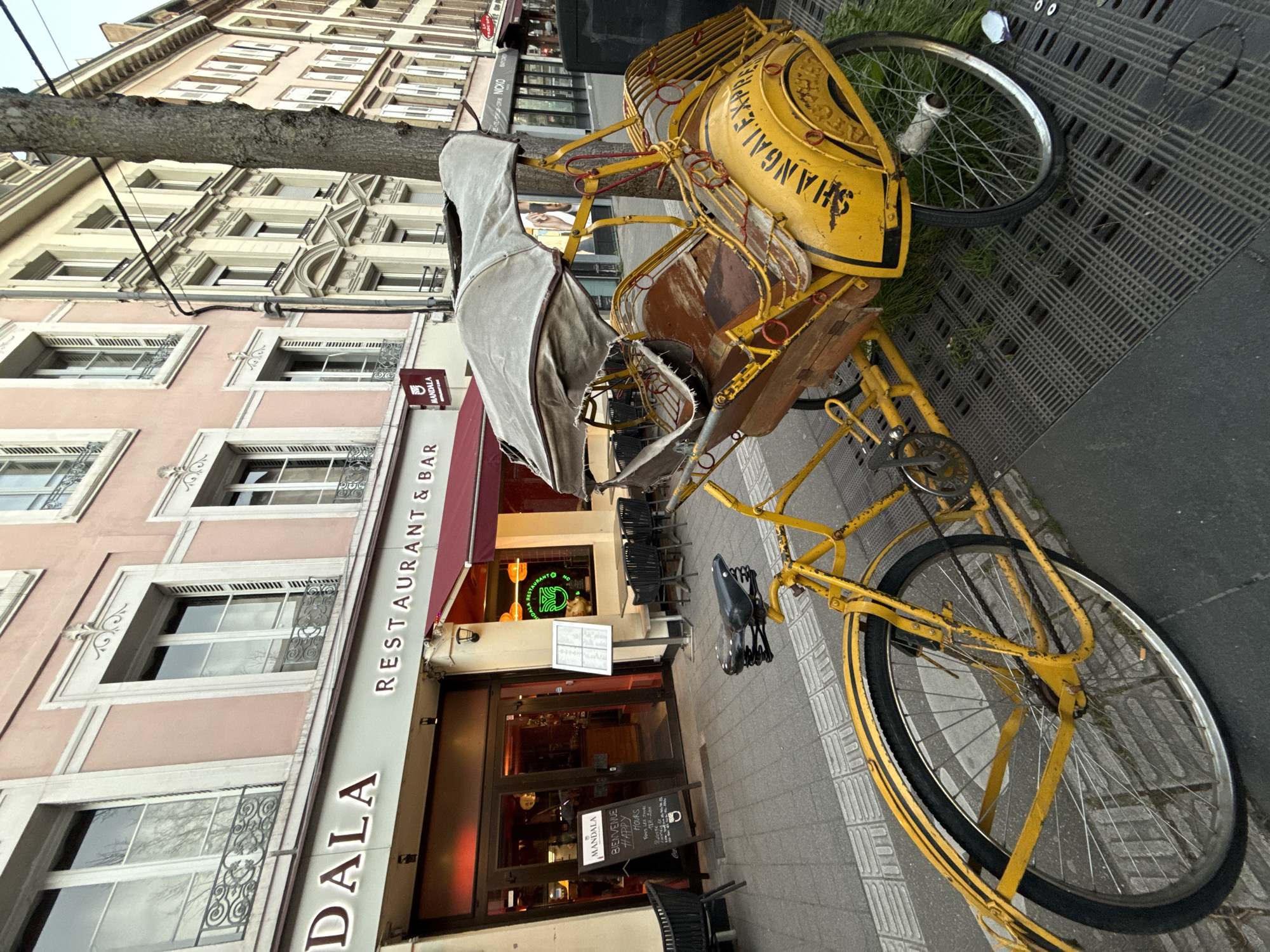 A heavily worn, bright yellow bicycle-powered pedicab, displaying "SHANGHAI EXPRESS" in black lettering on its rear fender, is parked on a paved sidewalk. The pedicab features two passenger seats with weathered wooden bases and torn upholstery, covered by a light grey canvas canopy. Its metal frame exhibits significant rust and peeling paint. The front wheel and pedals are visible, indicating it is a functioning vehicle despite its condition.
The pedicab is positioned next to a tree trunk and partially over a square metal grate integrated into the sidewalk. Behind it is the facade of "MANDALA RESTAURANT & BAR," a three-story building with a light pink painted facade and cream-colored architectural trim around its windows. The restaurant features large dark glass doors, a red awning, and an outdoor seating area with several black chairs and small tables. A neon sign depicting a green symbol is visible inside the restaurant. Two white menu stands are placed near the entrance.
Further down the street, to the right, a sequence of multi-story European-style buildings with light-colored facades and numerous windows, many with white shutters, lines the street. The sky is overcast. No individuals are present or visible within the scene.
Based on the visible signage for "MANDALA RESTAURANT & BAR," the location is identified as Rue du Vieux Marché aux Poissons in Strasbourg, France.
