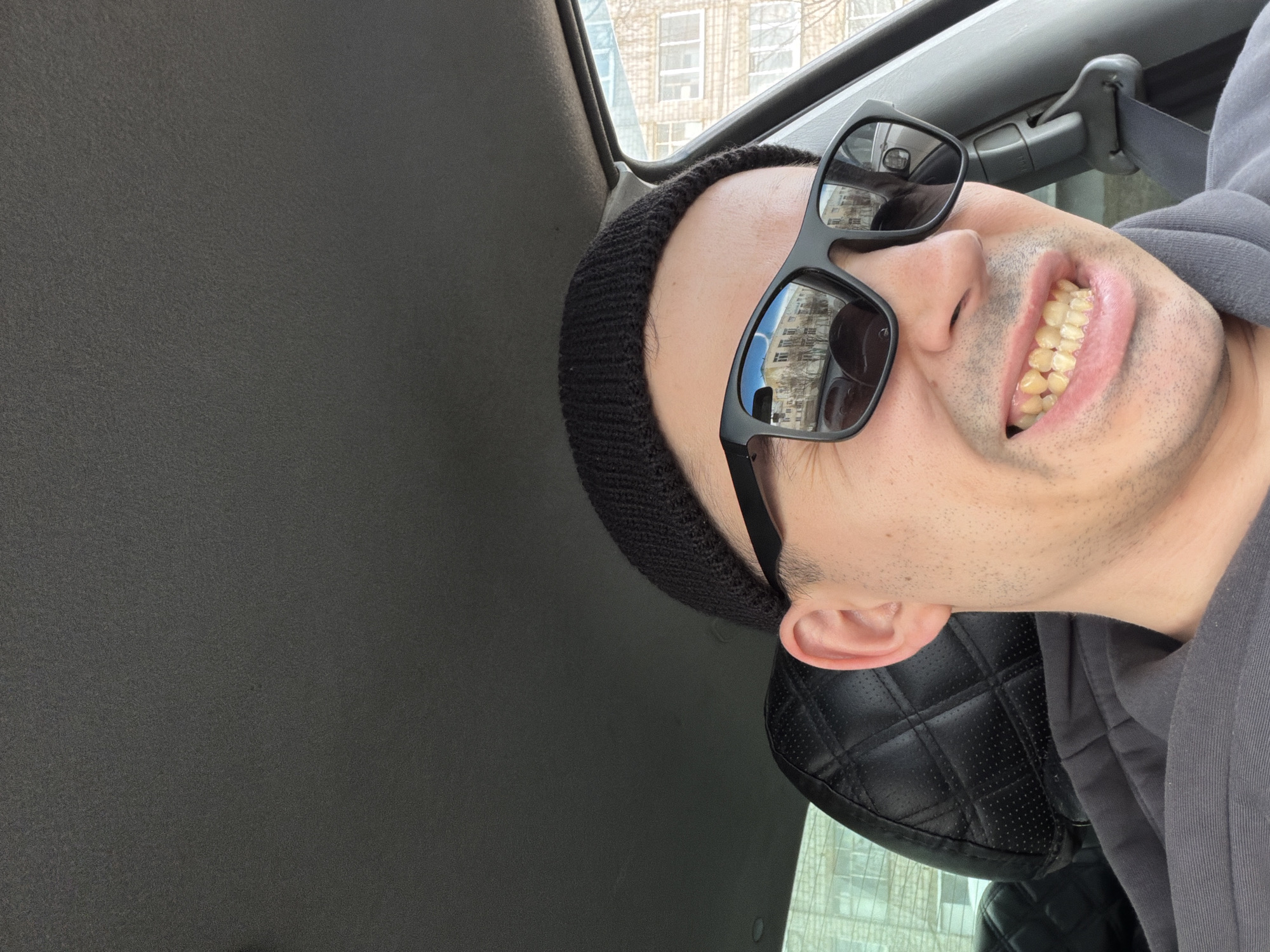 A selfie shows a person with a shaved head wearing a black beanie and sunglasses in a car. The reflection in the sunglasses shows a building with windows. Based on the architectural style in the reflection, this is potentially a location within the city of Izhevsk, Russia. The person is smiling, revealing their teeth. They are wearing a gray hoodie. The car's interior is visible, including the roof, seat, and seatbelt. The seat has a quilted pattern. The background is a blurry view from the car window.