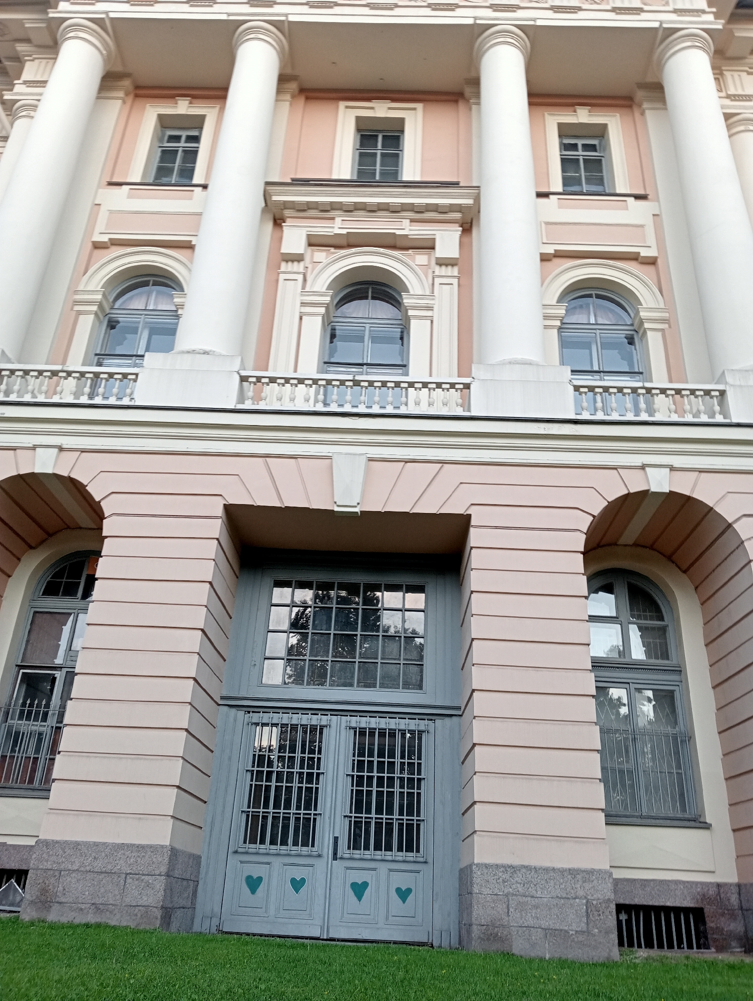 The image shows the facade of a large, classical building with prominent white columns. The building has a light pinkish-beige exterior with decorative moldings around the windows and arches.
The upper part of the building features several rectangular windows with dark frames. Below these, a balcony with a balustrade stretches across the facade, supported by decorative brackets. There are also arched windows on this level.
The lower part of the building is made of a similar pinkish-beige material, but with horizontal grooving. This section features larger arched openings, one of which contains a set of double doors painted a muted blue-grey color. These doors are secured with metal grilles and have three stylized green heart shapes painted on them, one on each door and one in the center. To the left and right of these doors are large, arched windows, also with dark frames and metal grilles. The lower base of the building is made of granite blocks.
The ground in front of the building is covered with green grass. The scene is lit by natural light, suggesting it was taken outdoors during the day. The perspective is from below, looking up at the building.
This building is identified as the Tsentral'nyy Muzykal'nyy Muzyey (Central Musical Museum), located at Italyanskaya Street, 22, in Saint Petersburg, Russia.