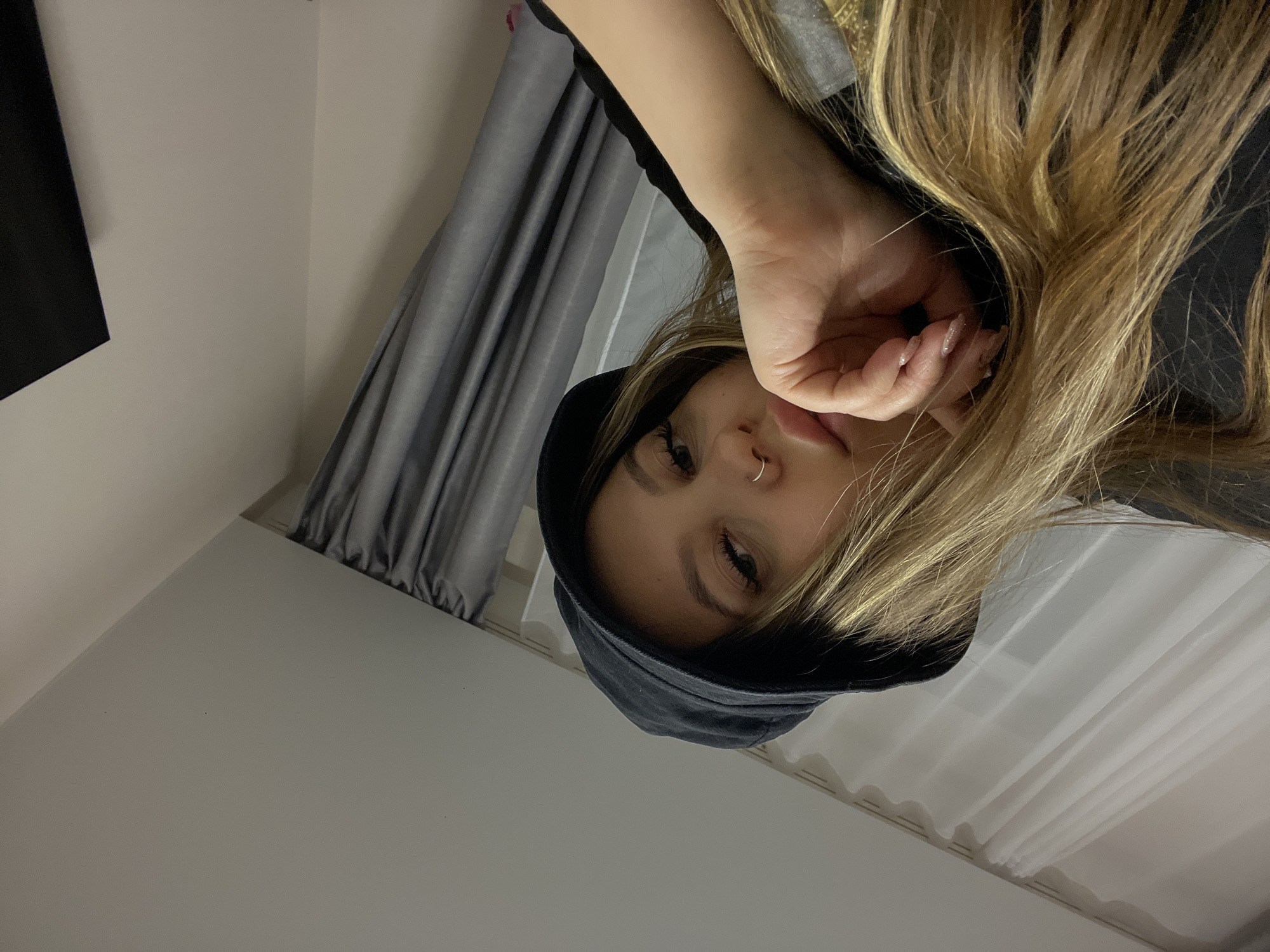 An indoor portrait features a young person, likely female, with long, blonde and light brown hair. The subject is wearing a dark hooded sweatshirt and has a silver nose ring on their right nostril. Their right hand, adorned with long, light-colored fingernails, rests near their mouth and chin area. The person looks directly at the camera with a neutral expression.
In the background, on the left side, are grey pleated curtains, with sheer white curtains visible behind them. The surrounding walls are plain and light-colored. The image is a close-up, likely a selfie, focusing on the person's upper body and face. The lighting is soft and even, suggesting indoor conditions. No visible text is present. The context provided indicates the location is Krasnodar, Russia.