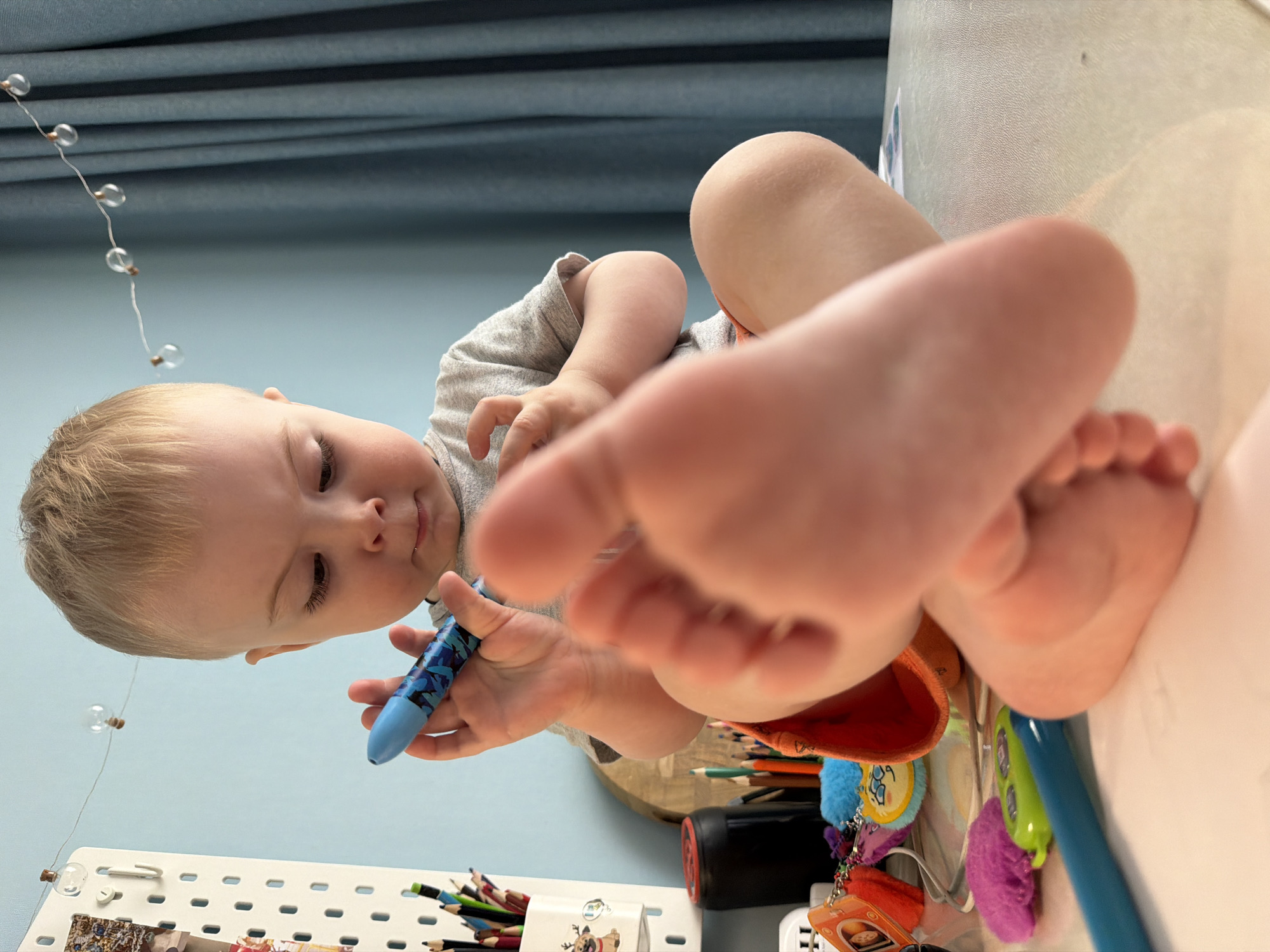 A young child with fair hair and a light gray shirt sits on a table with various items. In the child's hand is a blue marker. The child's feet are prominently displayed in the foreground. The table contains pencils, a red container, and several keychains. The background consists of a blue wall, string lights, and a blue curtain. The location is presumed to be a home environment in Votkinsk, Russia.