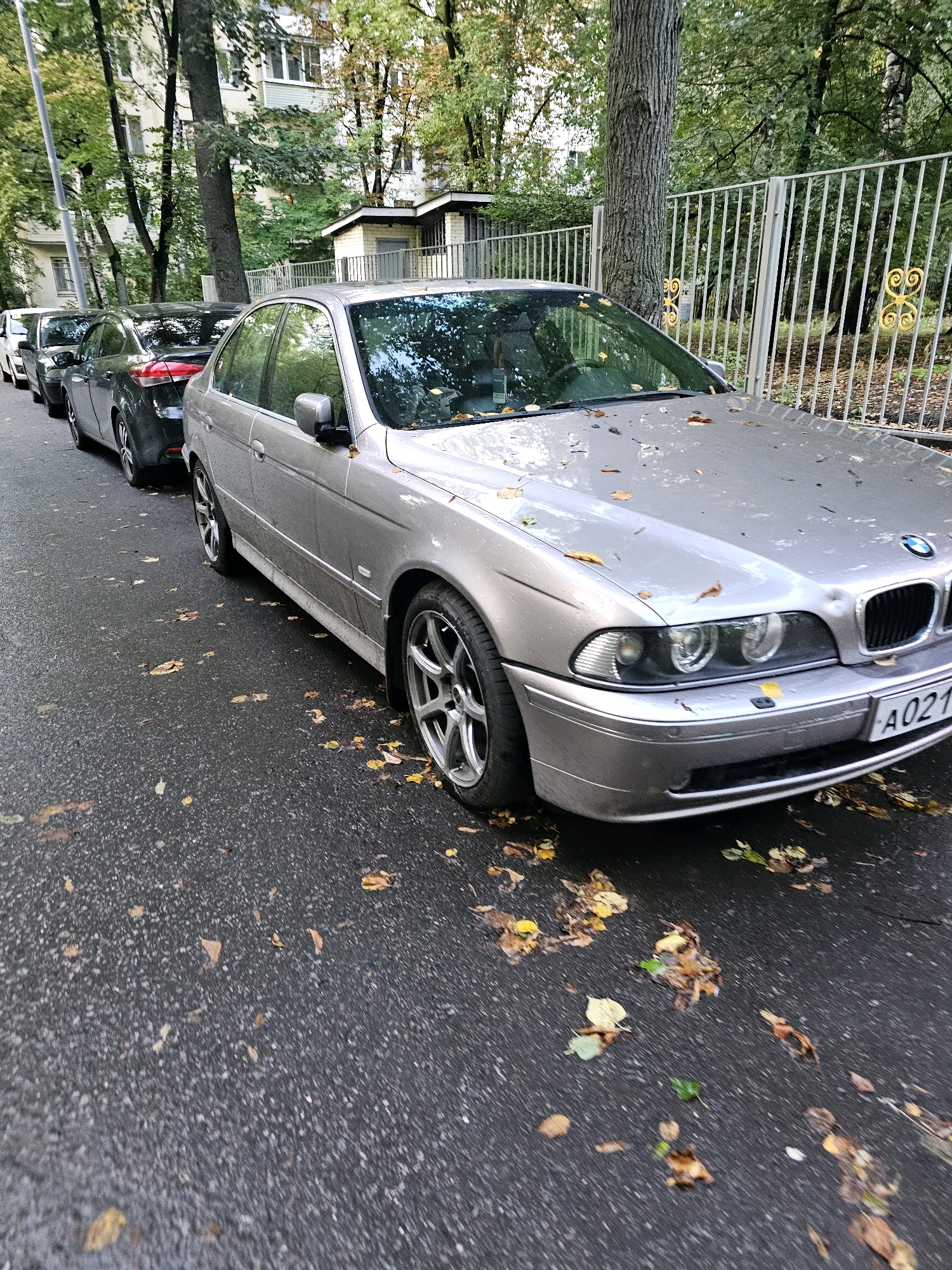 An overhead shot of a silver BMW 5 Series sedan parked on the side of a street in the city of Khimki, Russia. The car is facing right and is partially obscured by other cars parked in front of it. The street is wet and covered in fallen autumn leaves. A metal fence with decorative elements is visible behind the car. A building with windows and a small structure is in the background. The license plate of the BMW is visible and reads "A021".