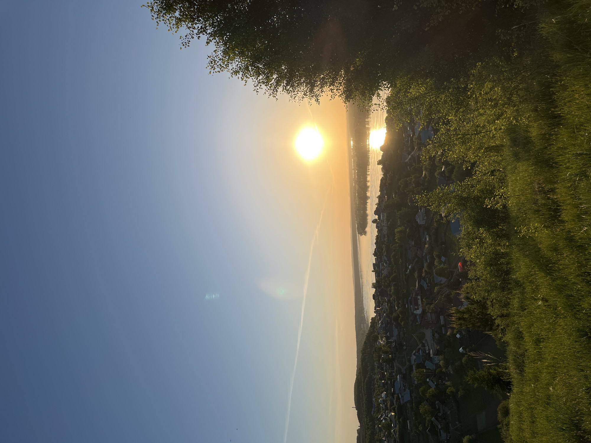 The image captures a sunny view of the city of Ольховка, Russia, likely during sunrise or sunset. The sun is prominently positioned near the horizon, reflecting on a body of water, presumably a river or lake. The surrounding area features a dense concentration of houses and buildings, characteristic of a residential area. A lush green landscape with trees and grass frames the urban sprawl, suggesting the scene is taken from a high vantage point on the outskirts of the city. A contrail stretches across the clear blue sky, indicating the passage of an aircraft. The photo is shot with the camera tilted, causing a slight distortion in the perspective of the scene.