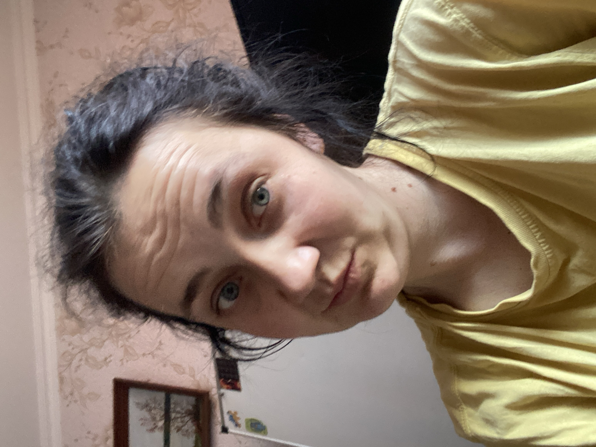 A woman with dark hair pulled back and light skin is looking at the camera with a slightly questioning expression. She is wearing a yellow t-shirt. Her face is in focus, while the background is slightly blurred. To the left of the woman, a white board or refrigerator with some colorful magnets is visible. Behind her, there is wallpaper with a floral pattern and a framed picture on the wall. The lighting appears to be indoor lighting. The overall impression is a casual, informal selfie taken indoors.