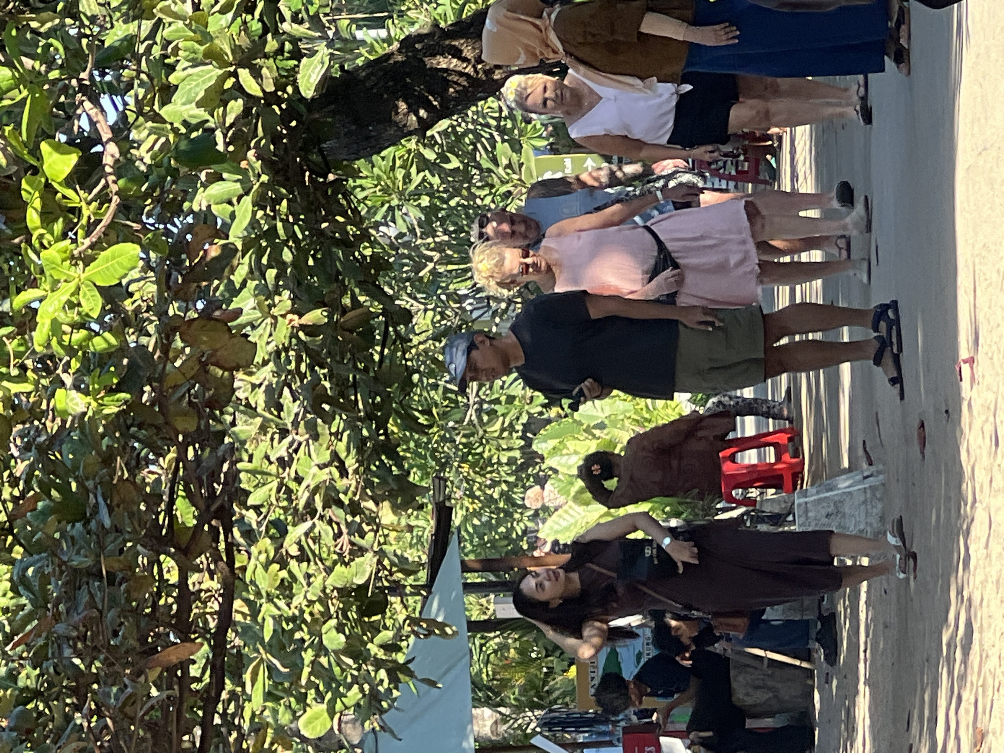A group of people are walking on a sandy path under the shade of trees. In the foreground, a woman in a dark brown dress and sandals is walking towards the right, touching her hair. To her right, a man in a dark t-shirt and khaki shorts is walking. Behind them, a woman in a pink dress with a black fanny pack and sunglasses is walking, followed by a man in a blue shirt and then two women in white tops. One of the women is wearing a light-colored headscarf and a brown top. In the background, there are market stalls with various items displayed, including what appears to be wooden carvings and textiles. There are also umbrellas providing shade. The scene suggests a marketplace or a busy street near a beach. The architecture and foliage are consistent with a tropical environment, potentially in Denpasar, Indonesia.