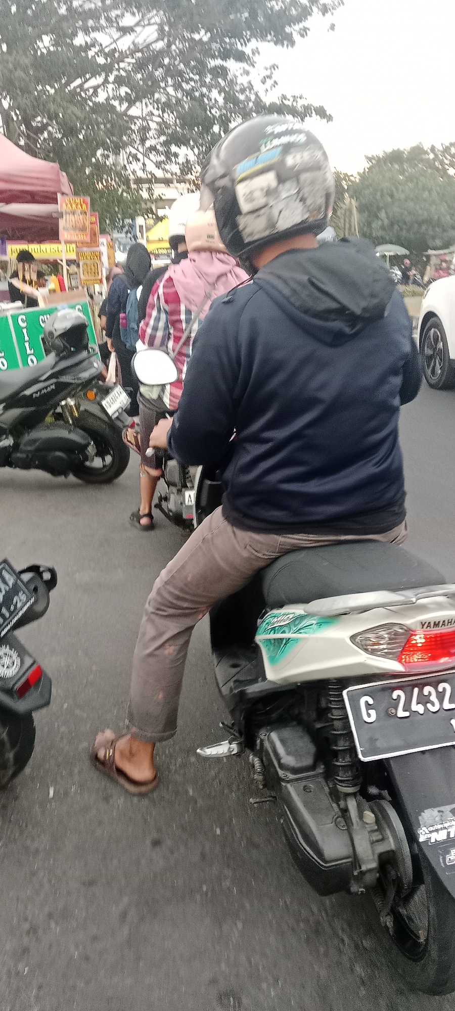 The image shows a street scene in Ngemplak, Indonesia, likely during daylight. In the foreground, a person wearing a dark blue hooded jacket and brown pants is riding a scooter. The scooter is white with green and blue graphics and has a license plate that reads "G 2432". The person is wearing a black helmet with decals. Behind this scooter, another person on a black scooter is visible, wearing a plaid shirt and a white bag over their shoulder, also with a helmet. Further in the background, there are several other scooters and motorcycles, indicating a traffic situation. Along the left side of the street, there are food stalls with red awnings and signage, some with text and images of food. Trees are visible in the background, and the sky appears overcast. The road surface is asphalt.