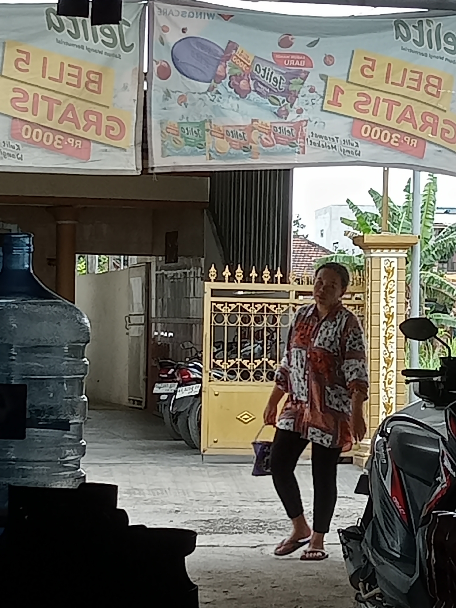 The image depicts a street scene in Simo, Indonesia, featuring a woman standing in front of a building. She is wearing a patterned shirt and dark leggings, with one hand holding a patterned bag and the other by her side. The woman is facing the camera and appears to be walking. In the background, there are motorcycles, a large water jug, and a gate. The scene is outdoors, with buildings and vegetation visible. A banner with Indonesian text is also prominently displayed.