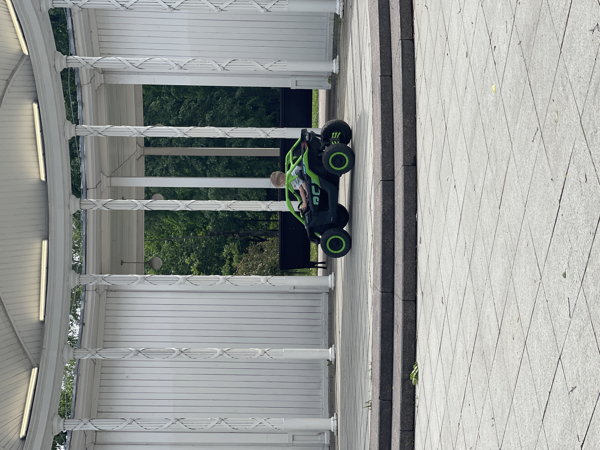 A medium-shot, outdoor image showing a child in a small, green and black electric car near a white colonnade structure. The child, wearing a light-colored shirt, is seated in the car, facing away from the viewer. The colonnade structure features white columns and a white wooden roof. The background shows green trees. The foreground is a paved area with a series of grey steps. The overall lighting suggests a sunny day.