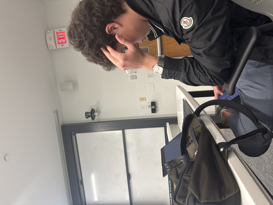 An indoor scene in a modern classroom or study environment in Wellesley, United States, captures a young adult male seated at a white desk. The male, positioned on the right side of the frame, has dark, curly hair and wears a black zippered jacket with a white and red circular Moncler logo on the left bicep, grey/blue trousers, and brown shoes. A silver-toned wristwatch is visible on his left wrist. His right hand is raised, fingers touching his forehead/temple area, as he looks downwards, possibly at a tablet or small device on his lap. On the white desk in front of him, a black laptop is open with a lit screen displaying text. A dark green/brown backpack rests to the left of the laptop. The background features a large white projection screen or whiteboard with a dark grey frame on the left. A black security camera or sensor is mounted on the white wall above waist height. An illuminated red "EXIT" sign is affixed to the ceiling in the upper right. White wall-mounted speakers and various electrical outlets are also present on the white wall. A portion of a wooden door frame is visible on the far right.