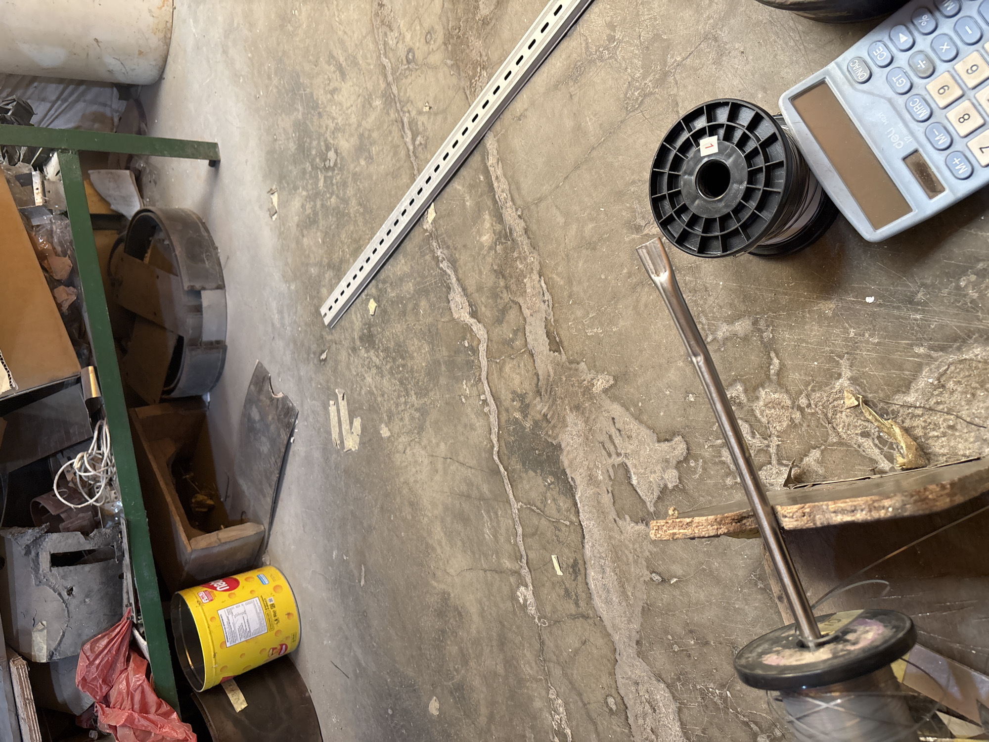 The image depicts a cluttered concrete floor in what appears to be a storage or workshop area. In the foreground, a metal rod is inserted through a spool of wire. To the right of the wire spool, a light blue calculator is placed on the floor. In the middle ground, a perforated metal strip lies diagonally across the concrete. In the background, a green metal shelf unit is visible, laden with various items including a yellow cylindrical container, a red plastic bag, and other debris. The overall environment suggests disarray and a lack of organization. There is no indication of the specific location within Telagamurni, Indonesia, nor are there any people or interactions depicted.