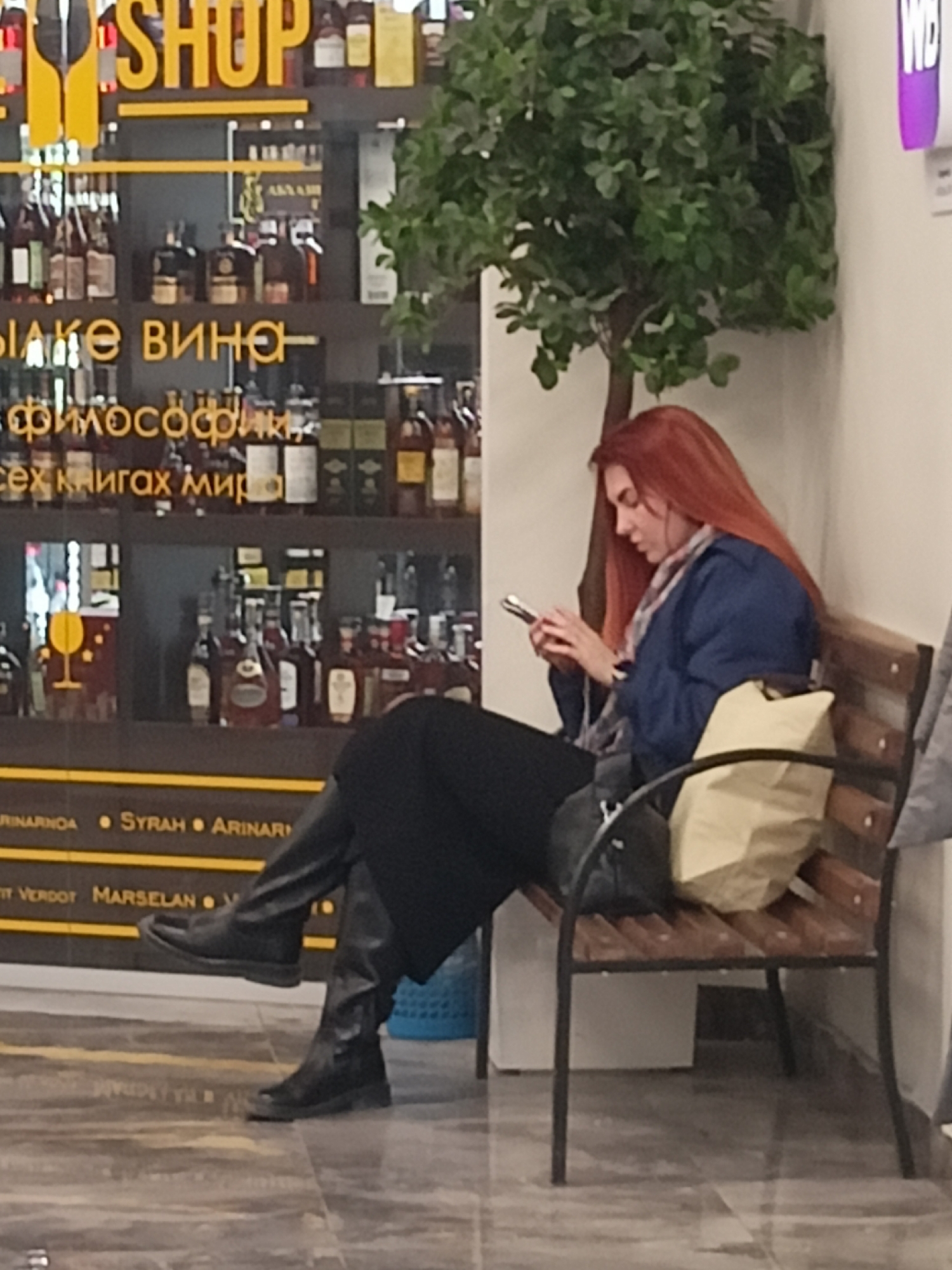 The image captures a woman with long red hair sitting on a wooden bench in what appears to be a shopping area in Novosibirsk, Russia. The woman is looking at her mobile phone. She is wearing a blue jacket, a scarf, a long black skirt, and black boots. A tan-colored tote bag sits next to her on the bench. Adjacent to the woman is a small potted tree. Behind her is a shop window displaying numerous bottles of alcohol and a sign that says "SHOP." Underneath the shop name, there are phrases in Russian, presumably indicating the type of alcoholic beverages offered. The flooring is a greyish-brown marbled pattern. A piece of a sign is visible in the upper right corner.