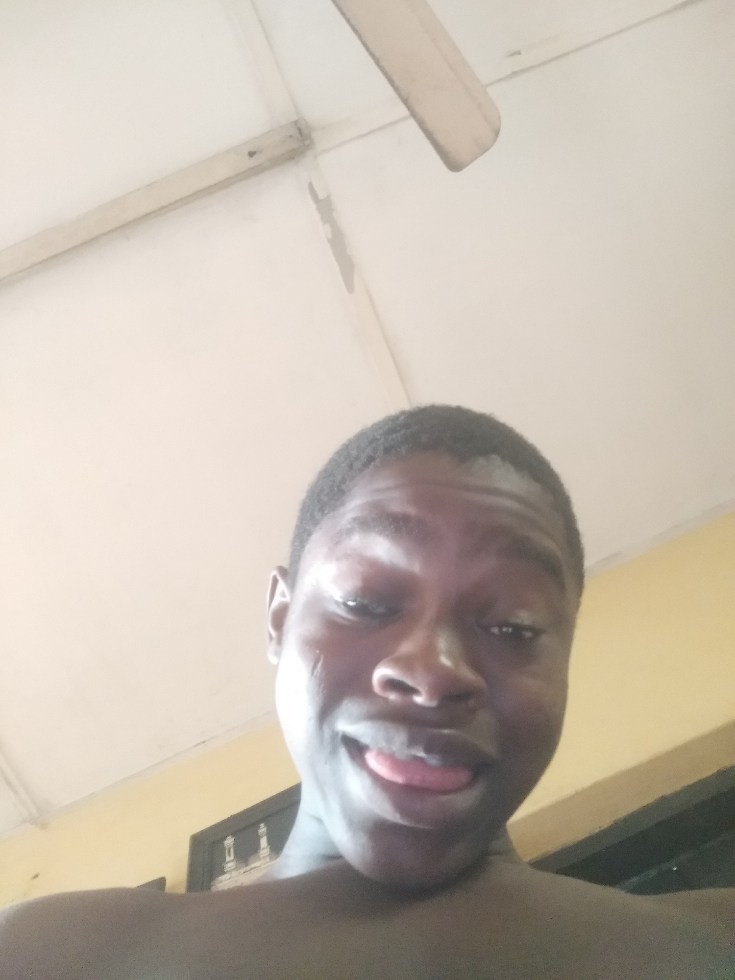 A close-up, angled shot of a young black man indoors. He is partially nude and sticking his tongue out. Above him is a white ceiling with a ceiling fan. The background shows a cream-colored wall and a partially visible framed picture. The location, Shangisha, Nigeria is not evident from the image.