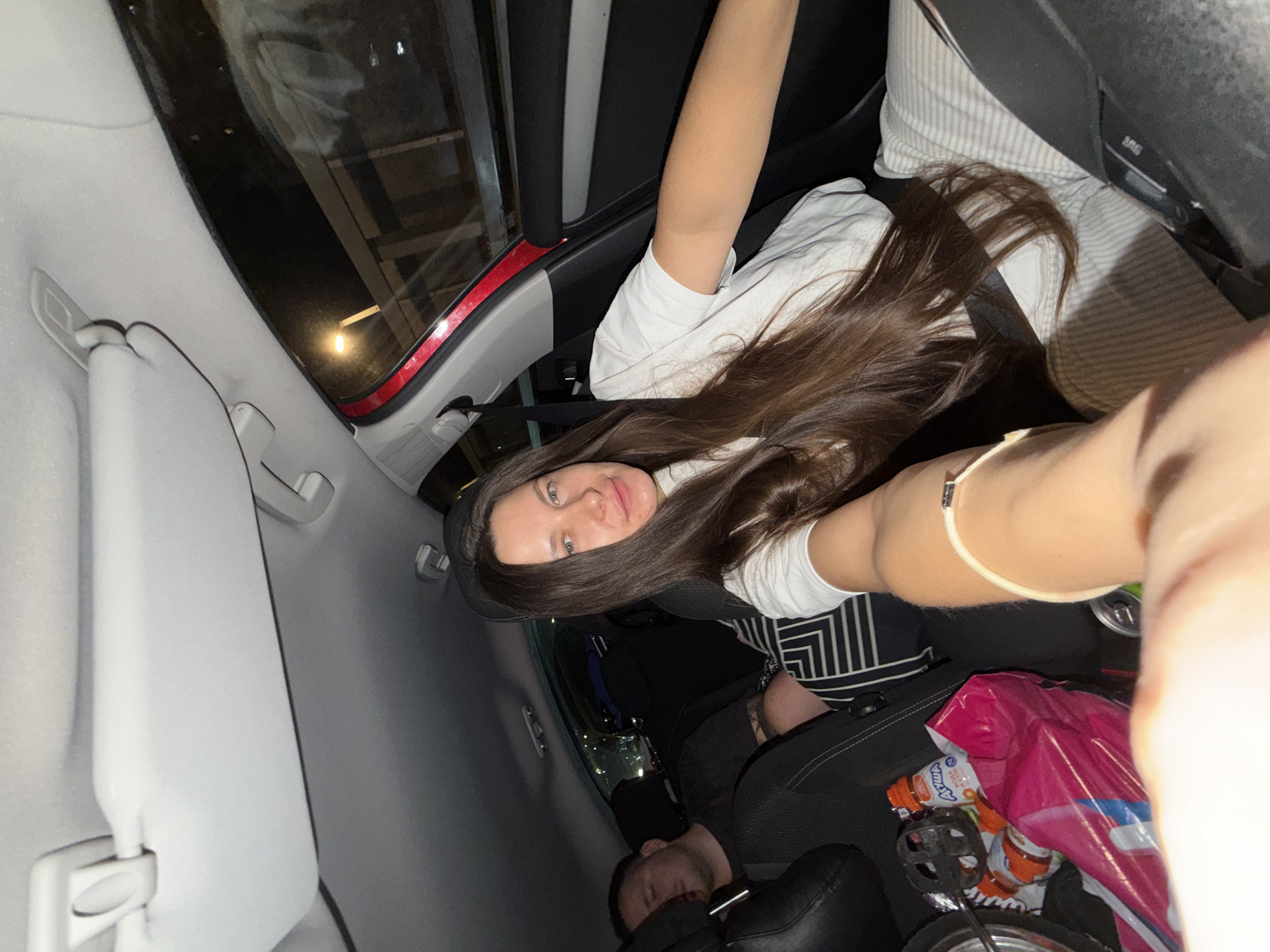 A long-haired woman in a white t-shirt and striped pants is taking a selfie inside a car at night. She is in the passenger seat, with her arm extended towards the camera. A man is asleep in the back seat, wearing a black t-shirt. There are various items in the car, including bags, bottles, and what appears to be a bicycle pedal. The scene is dimly lit, with the car interior illuminated by ambient light from outside. The background suggests a city environment, possibly Moscow, Russia, given the presence of streetlights and buildings visible through the car windows.