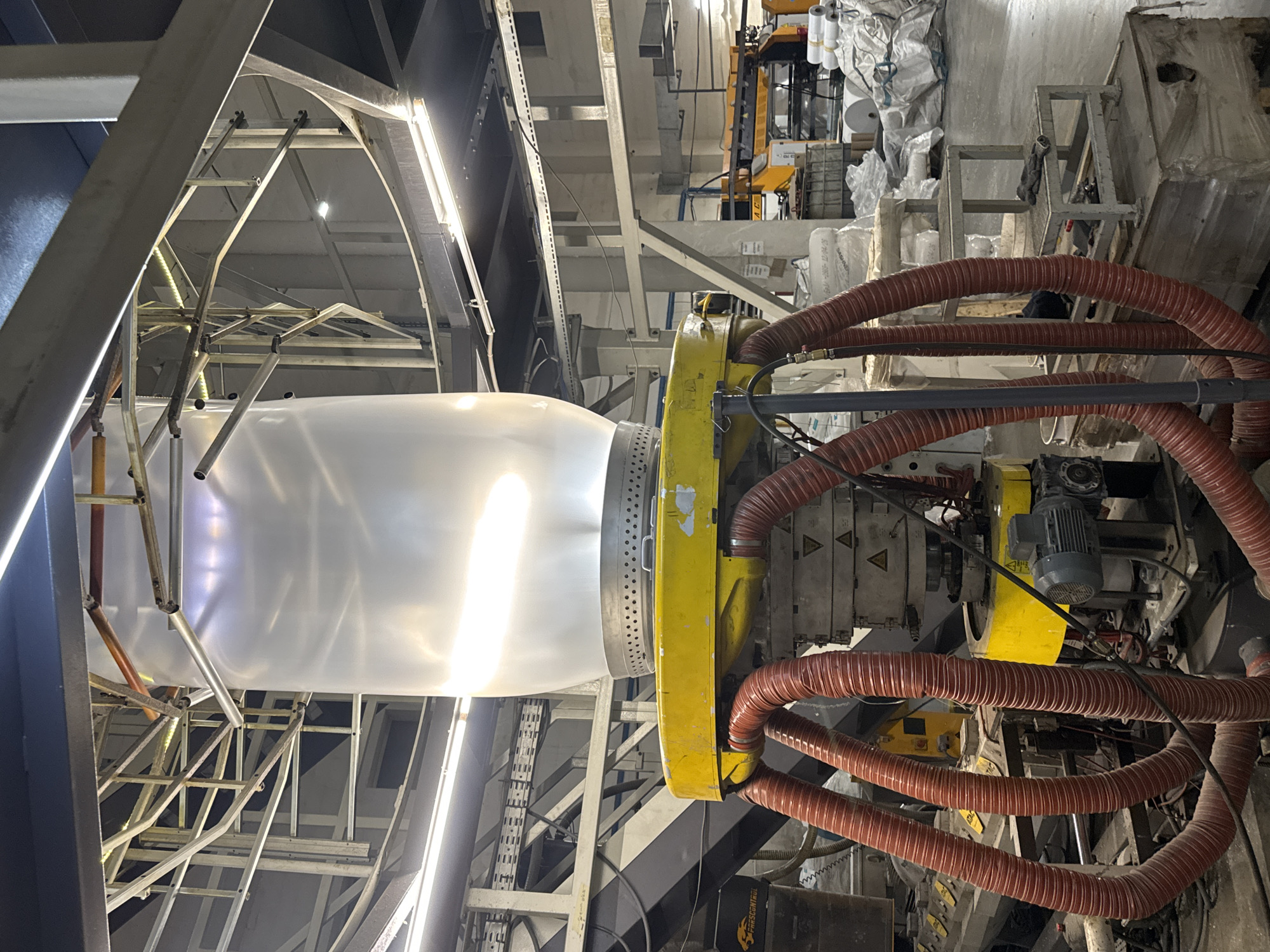 The image shows industrial equipment, specifically a blown film extrusion machine, likely in a factory setting. A large, translucent plastic bubble is being formed, held up by a metal frame structure. A yellow machine component is visible with multiple orange, flexible hoses connected to it. There is an electric motor and various control panels. The background reveals a cluttered industrial environment with metal beams, support structures, and stacks of materials, potentially rolls of plastic film. The location of the image is in the city of Perm, Russia. There are no people visible in the image.