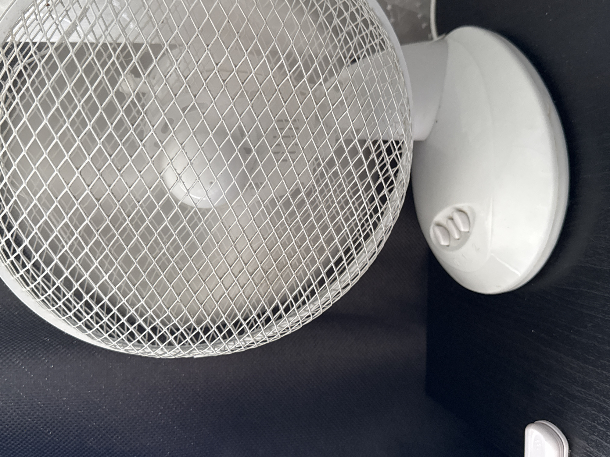 This is a close-up, eye-level shot of a white electric fan with a wire mesh grill. The fan is positioned on a dark, textured surface, likely a desk or shelf. Behind the fan, a dark, fabric-like background is visible. The fan's base has three control buttons labeled '1', '2', and '3'. A small, white object, possibly a cap or cover, is also visible on the surface to the left of the fan. The lighting is neutral, casting soft shadows. The scene is indoors, and there are no people present. The location is not specified within the image or its metadata.