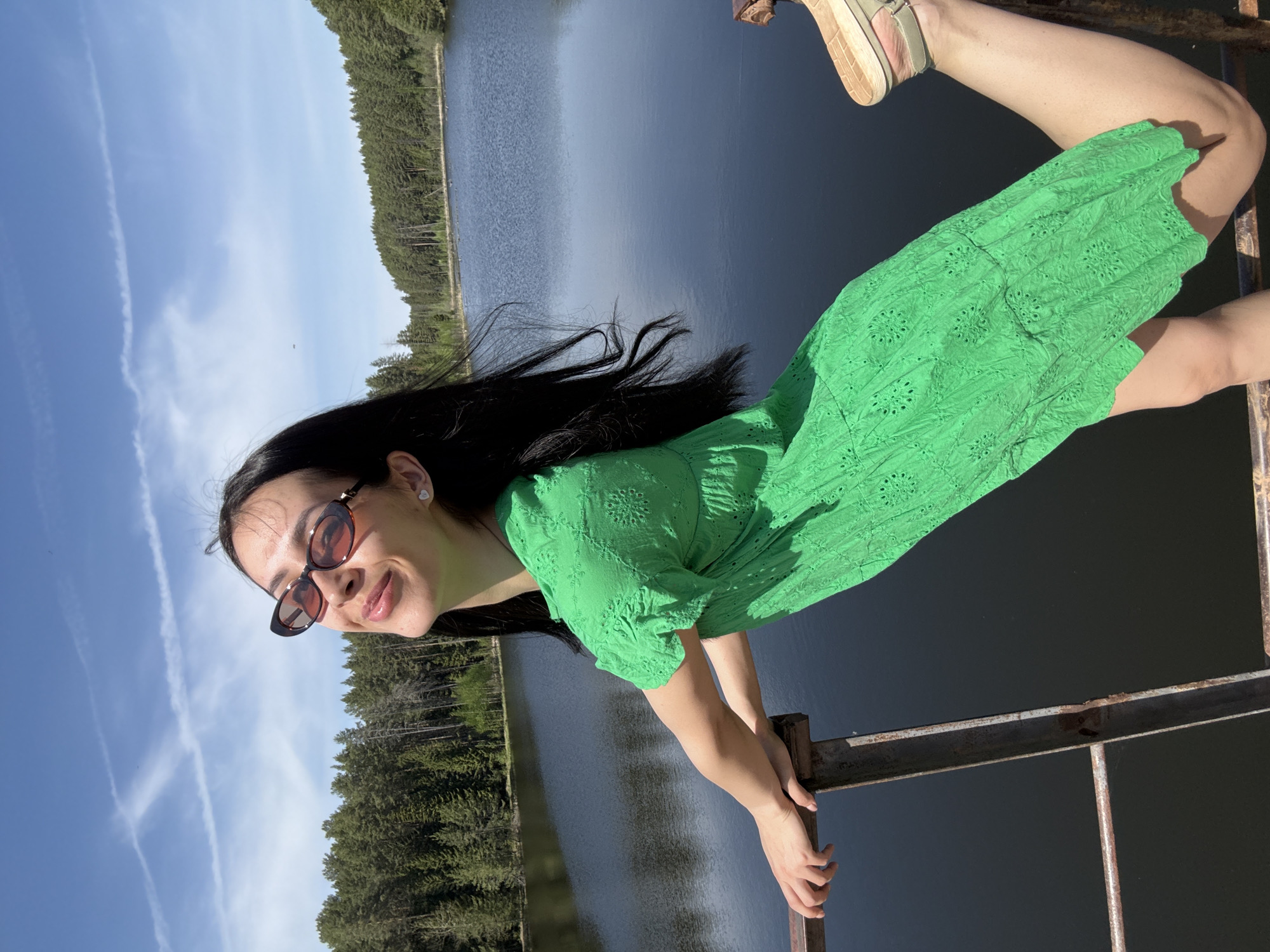 A woman is holding onto a rusty metal structure. She wears a green dress, sunglasses, and sandals. Her left leg is raised behind her. Behind the structure is a lake surrounded by trees. The sky is blue with white clouds. This is likely a bridge or overlook at a lake in Votkinsk, Russia.