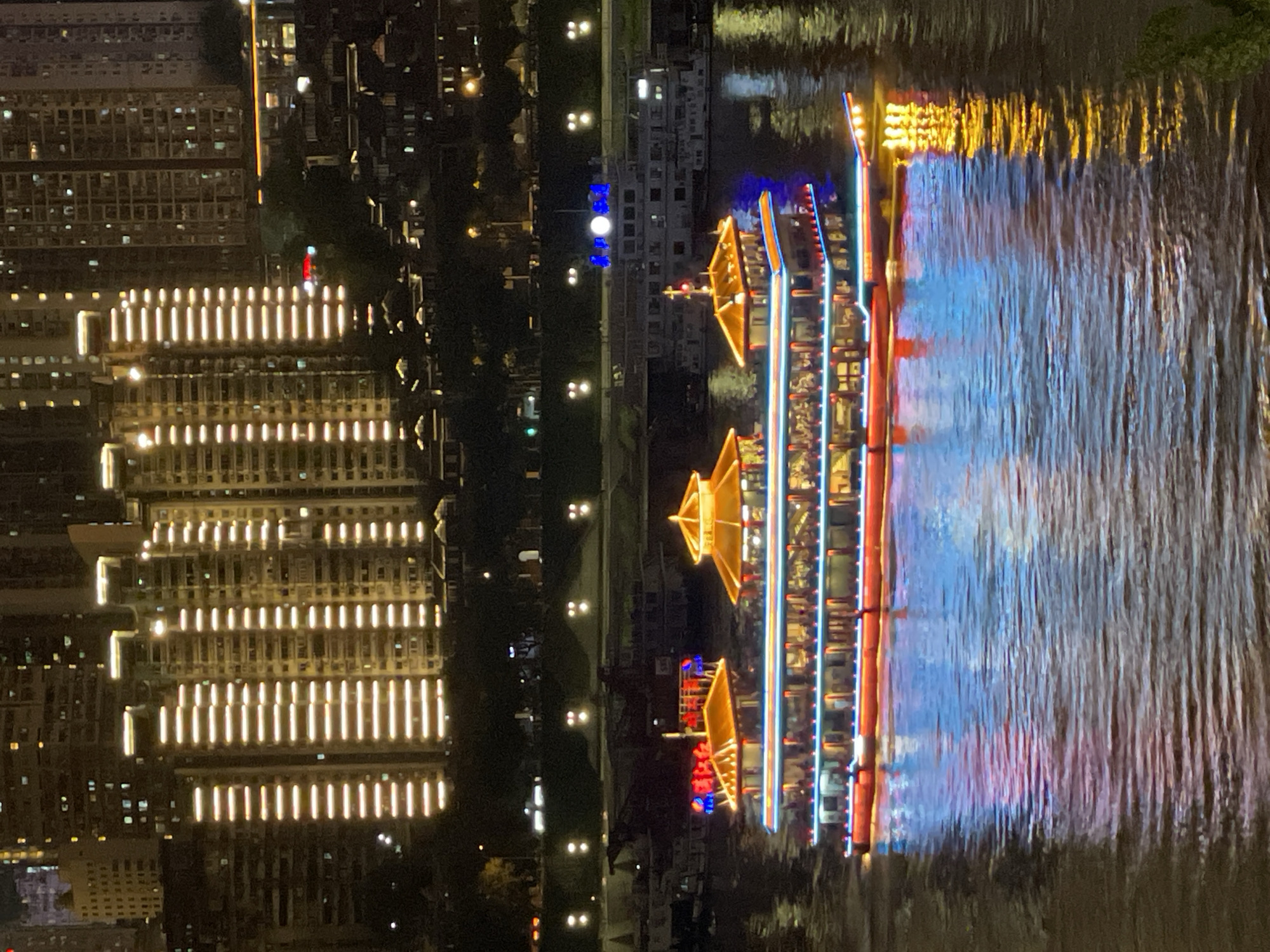 This is a night-time, eye-level photograph taken from a body of water, likely a river, looking towards a city skyline and a boat. In the foreground, the water reflects the colorful lights of the boat and the city. The main subject is a large, multi-decked boat, illuminated with bright red, blue, and yellow lights, traversing the water from left to right. The boat appears to be carrying passengers on its upper decks, visible as silhouettes against the internal lighting.
In the middle ground, there are other boats and structures along the riverbank, also lit up. The background is dominated by a dense collection of tall, modern buildings, illuminated with rows of warm yellow lights, creating a striking vertical pattern against the dark night sky. The buildings are clustered together, suggesting a high-density urban environment.
The overall scene depicts a vibrant night view of a city, emphasizing the interplay of artificial lights on the water, the architecture, and the passing boat. The location is identified as Nan'an District, China, which is known for its prominent riverfront development and illuminated skyline.