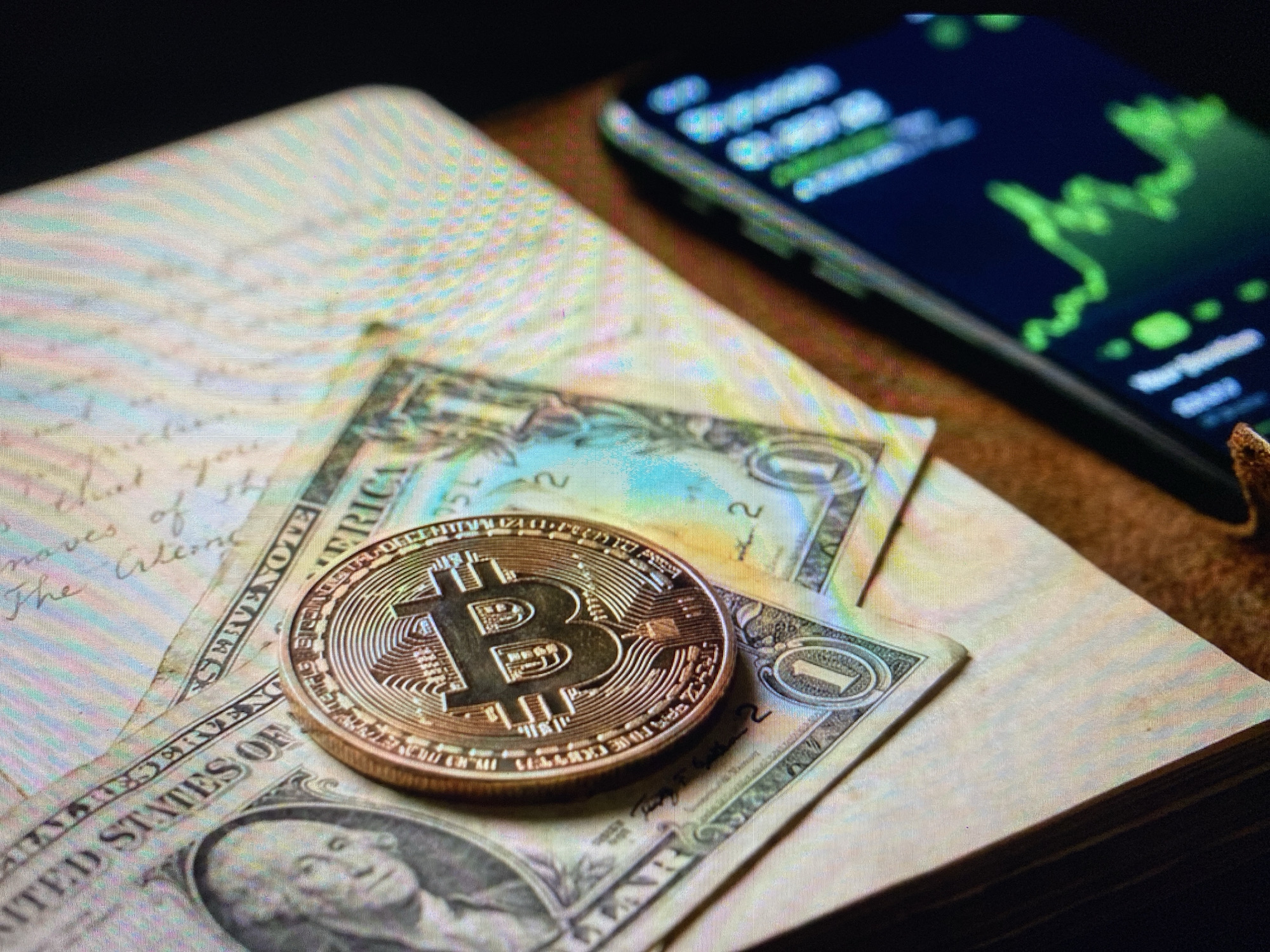 A close-up shot shows a Bitcoin coin and two US dollar bills placed on top of an open notebook with handwritten text. The notebook pages are light-colored and have visible lines of text. The dollar bills are positioned diagonally, with one bill partially obscuring the other. The Bitcoin coin, a replica with the iconic "B" symbol, is centrally placed on the dollar bills. In the background, a smartphone displays a green line graph, likely representing financial data. The overall setting appears to be indoors, with a dark background and warm lighting, emphasizing the metallic sheen of the Bitcoin and the paper texture of the currency. There is no visible information in the image to suggest a location in Kuta Selatan, Indonesia.